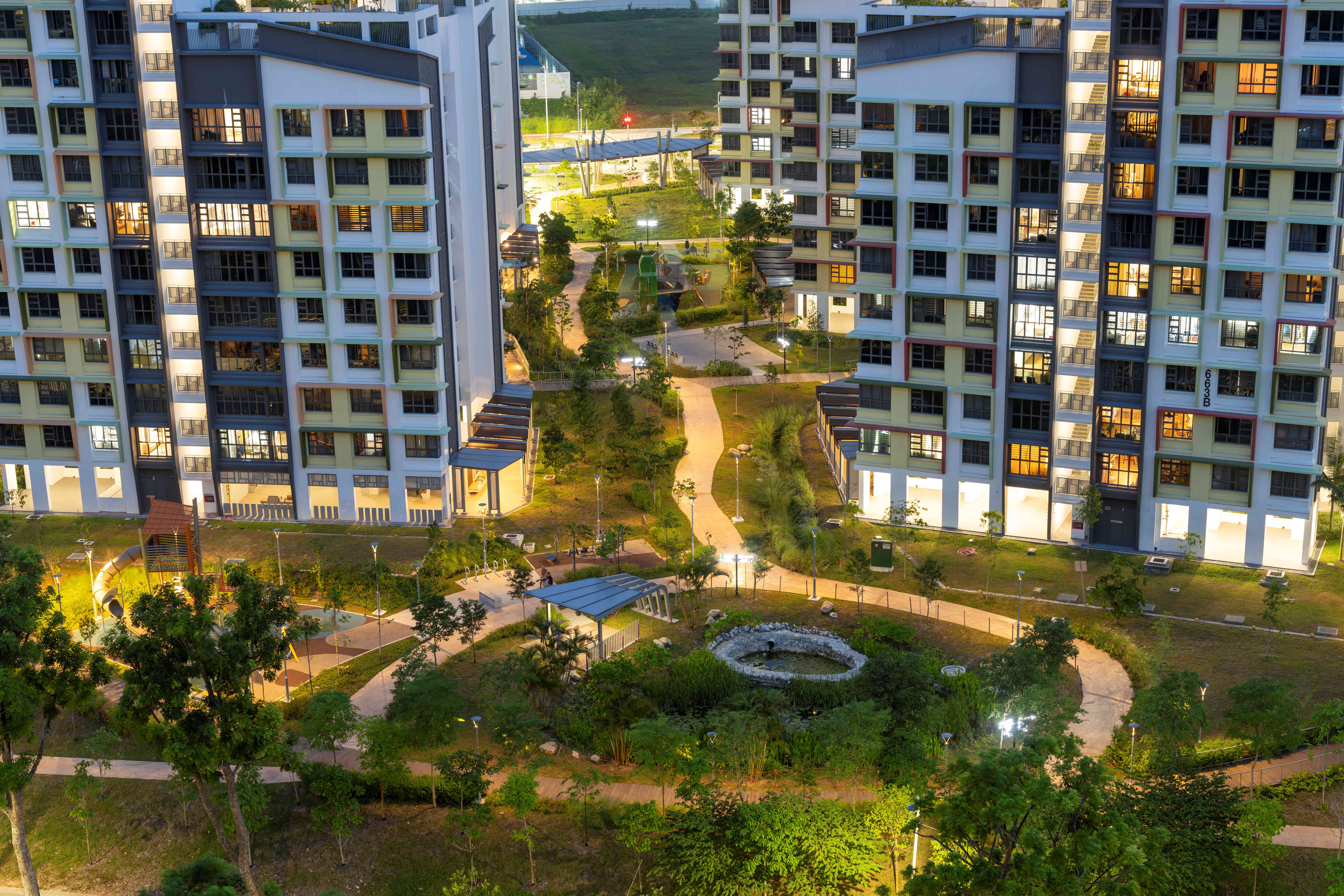 Courtyard between tall apartment buildings, with lit windows, winding paths, trees, playground, and a pond.