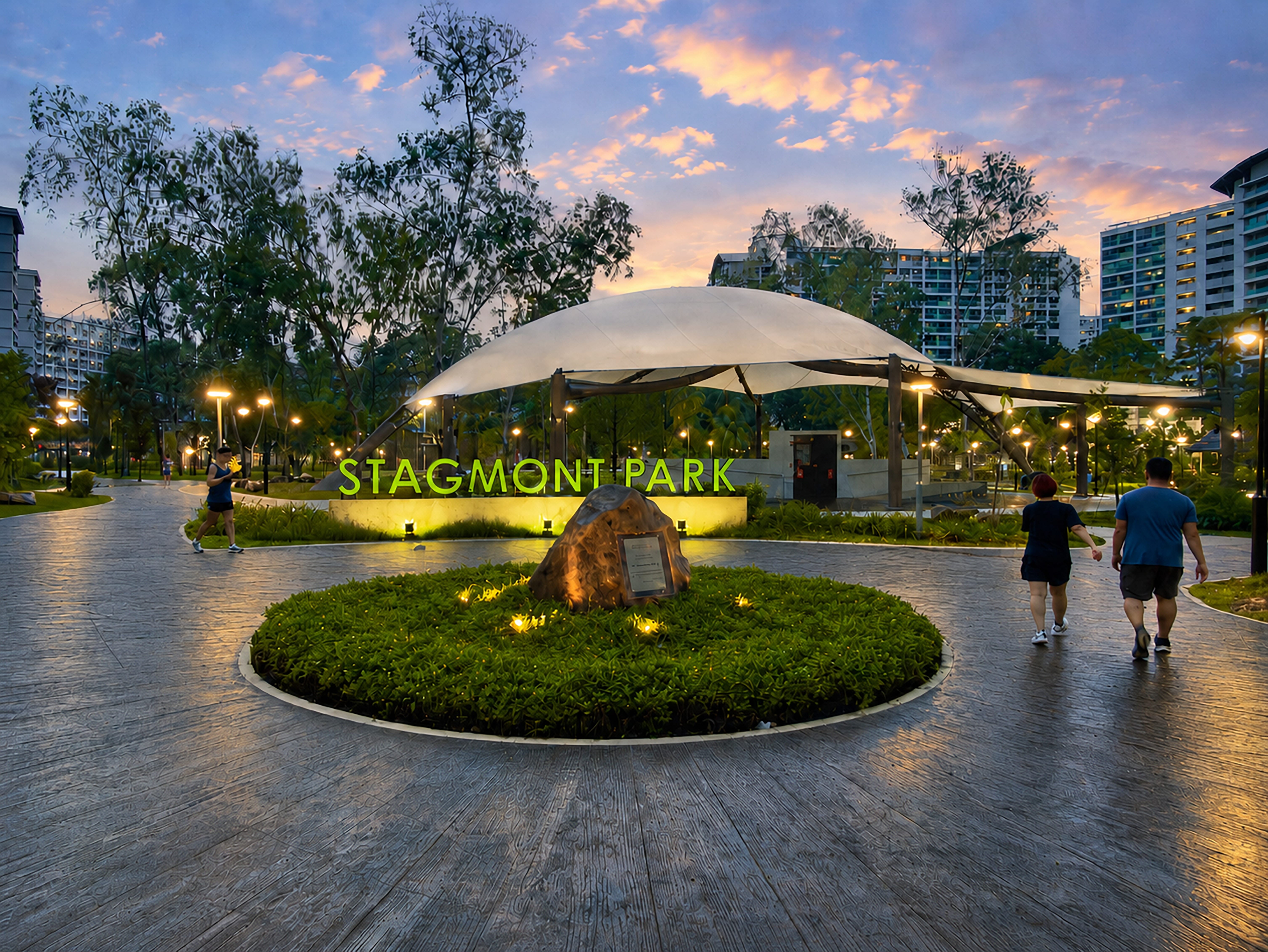 Dusk view of Stagmont Park entrance with lit sign, canopy pavilion, circular garden, and walkers on paths