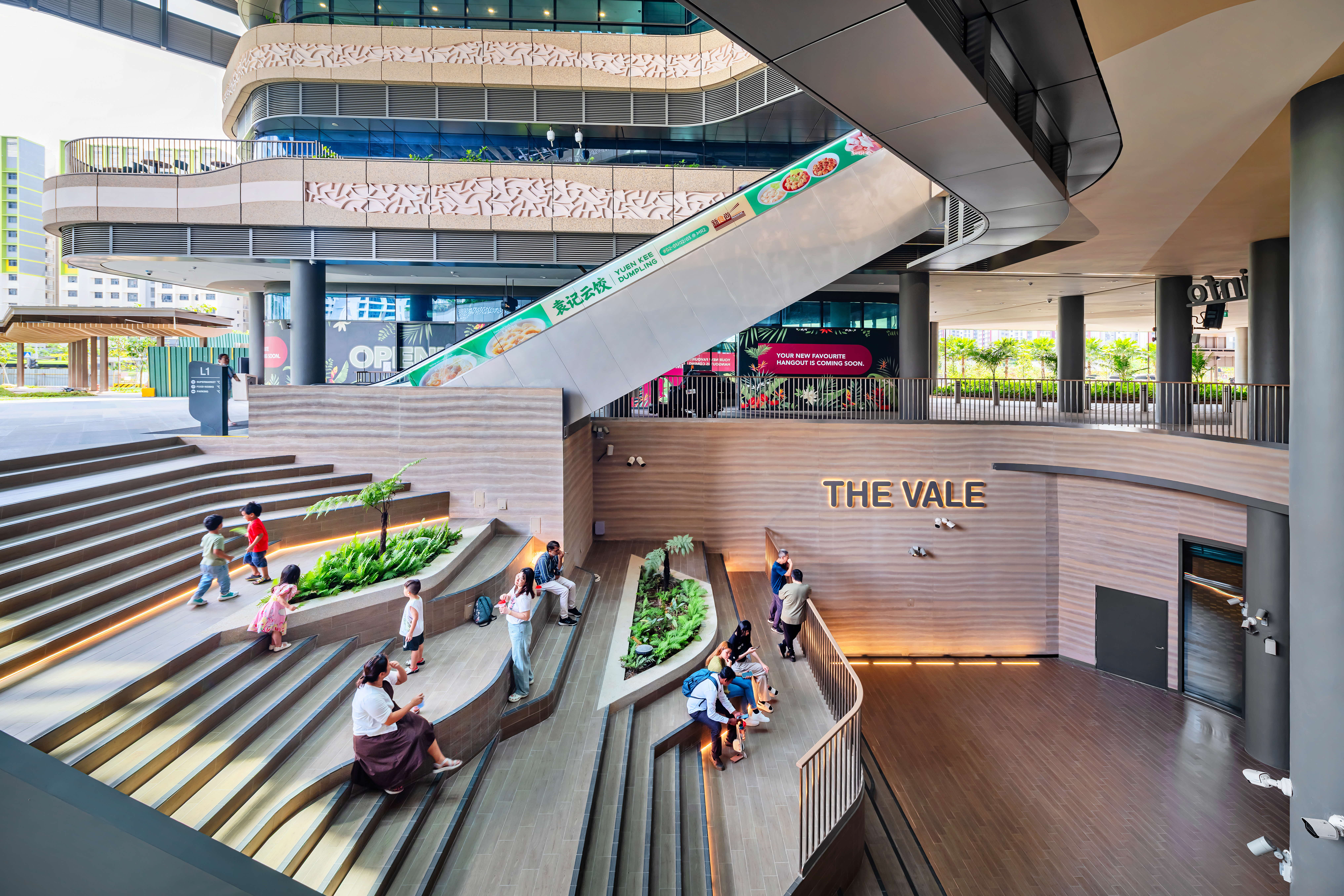 Open-air mall atrium with tiered amphitheater steps, people sitting, and escalator above The Vale sign