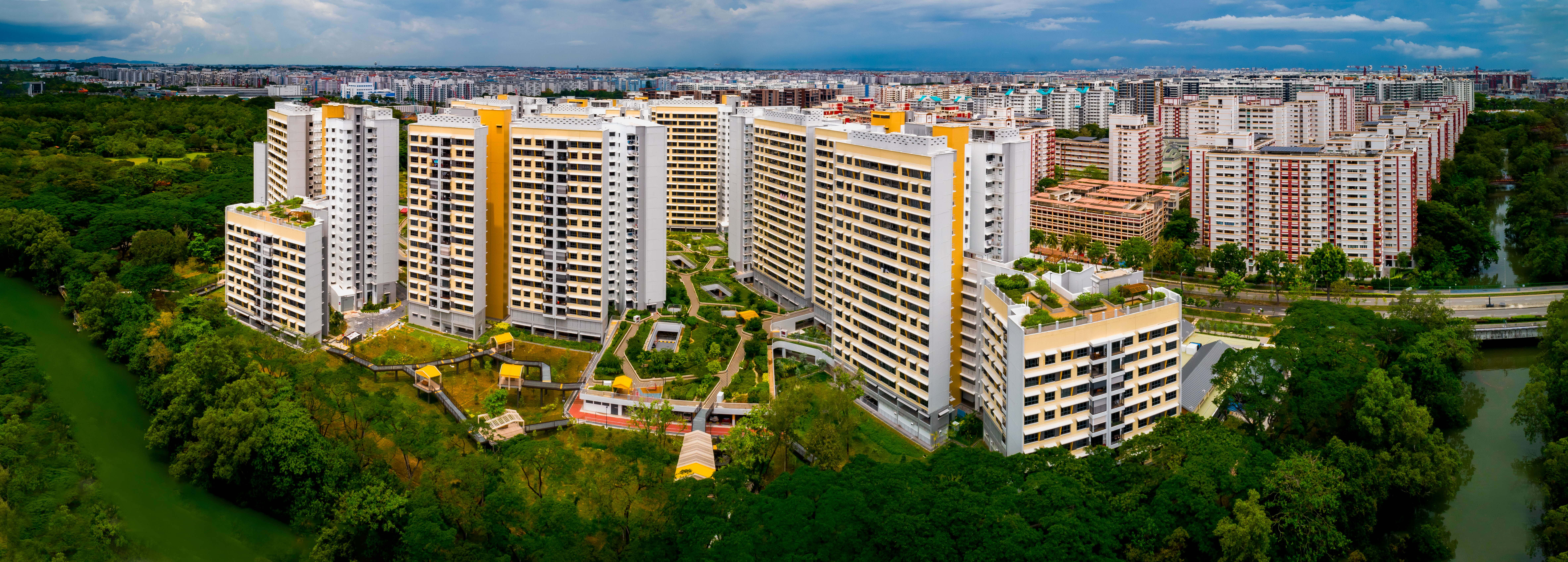 Aerial view of tall apartment blocks with courtyards, surrounded by dense greenery and a canal under clouds