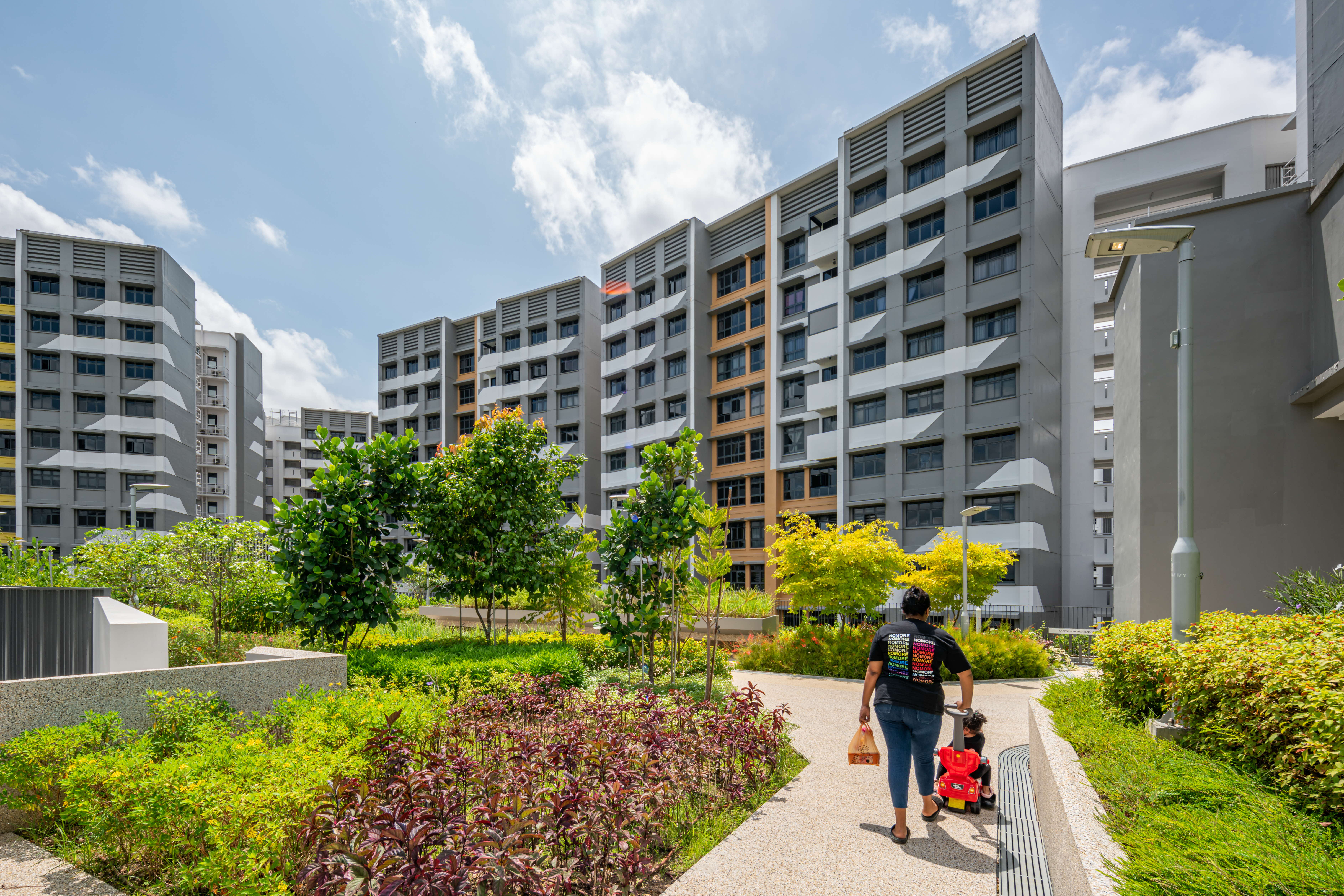 Person and child walk on a path through a garden courtyard between tall gray apartment buildings.