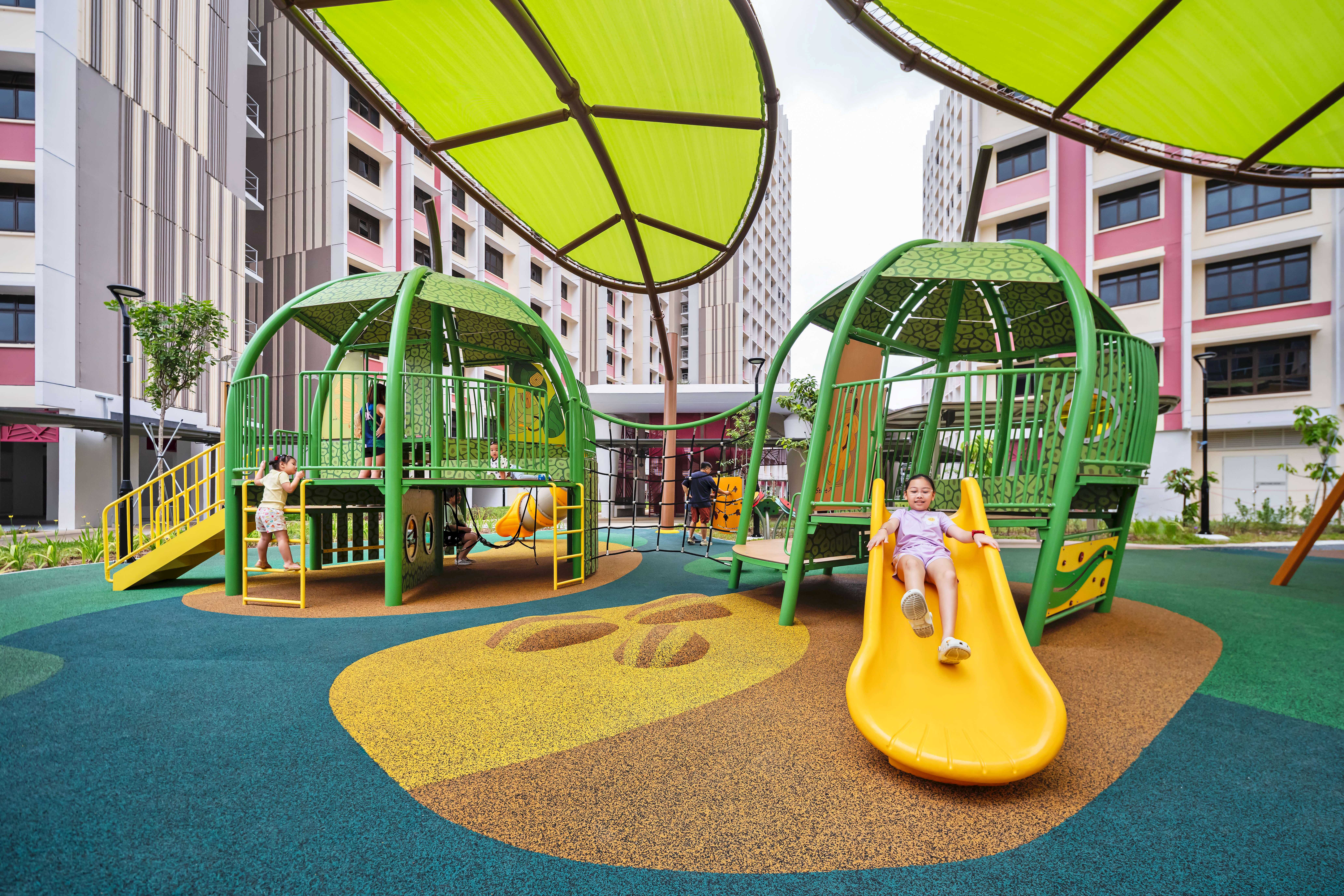 Children playing on green playground structures, one sliding down a yellow slide between apartment buildings.