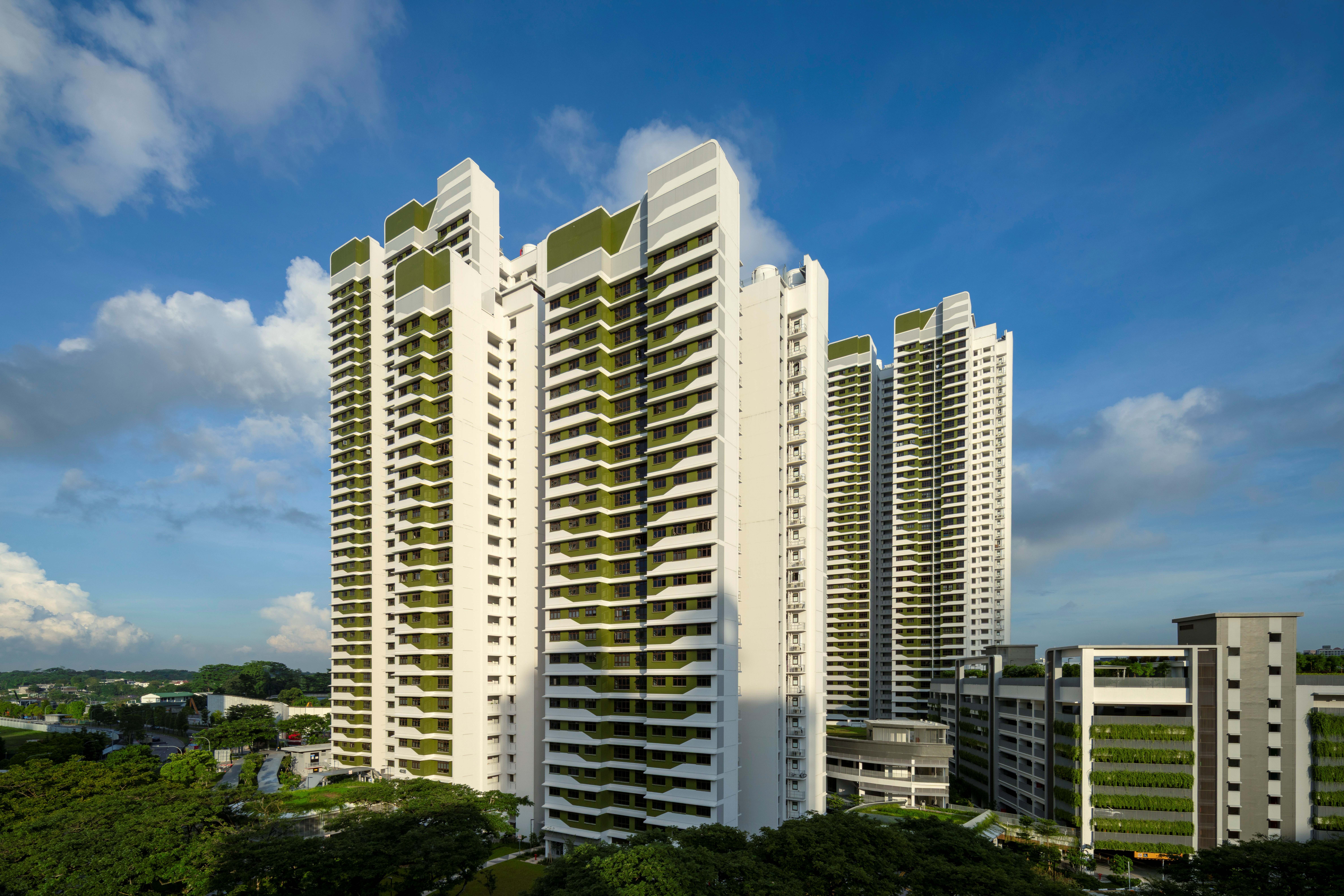 Three tall white apartment towers with green accents rise above trees under a bright blue sky with clouds.