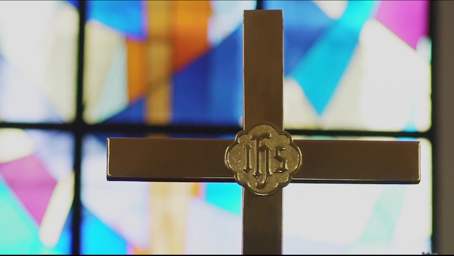 A gold metal cross with an IHS medallion at its center, set against colorful stained glass windows.