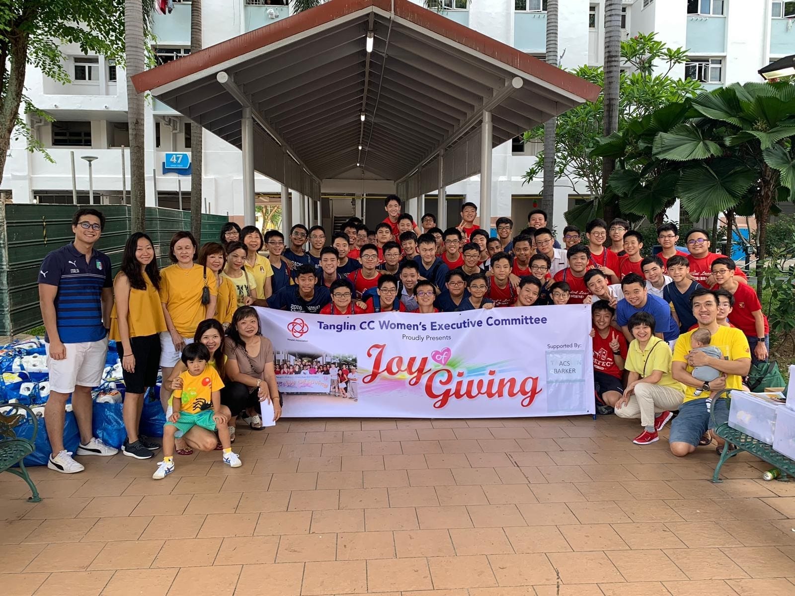 Large group posing outdoors with a banner reading Tanglin CC Women's Executive Committee Joy of Giving.