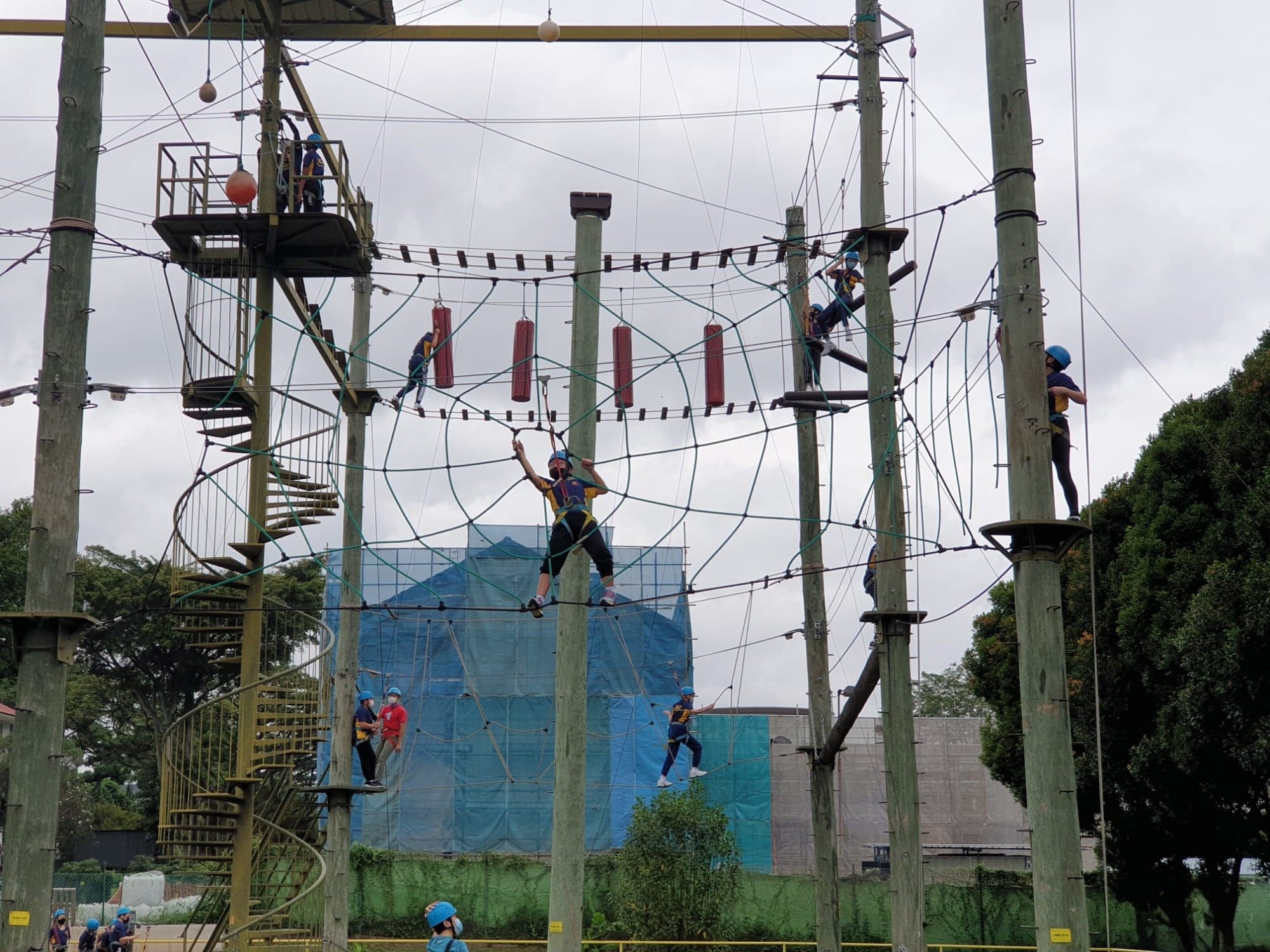 Students wearing helmets navigate a tall outdoor high ropes course with nets, cables, and wooden poles.