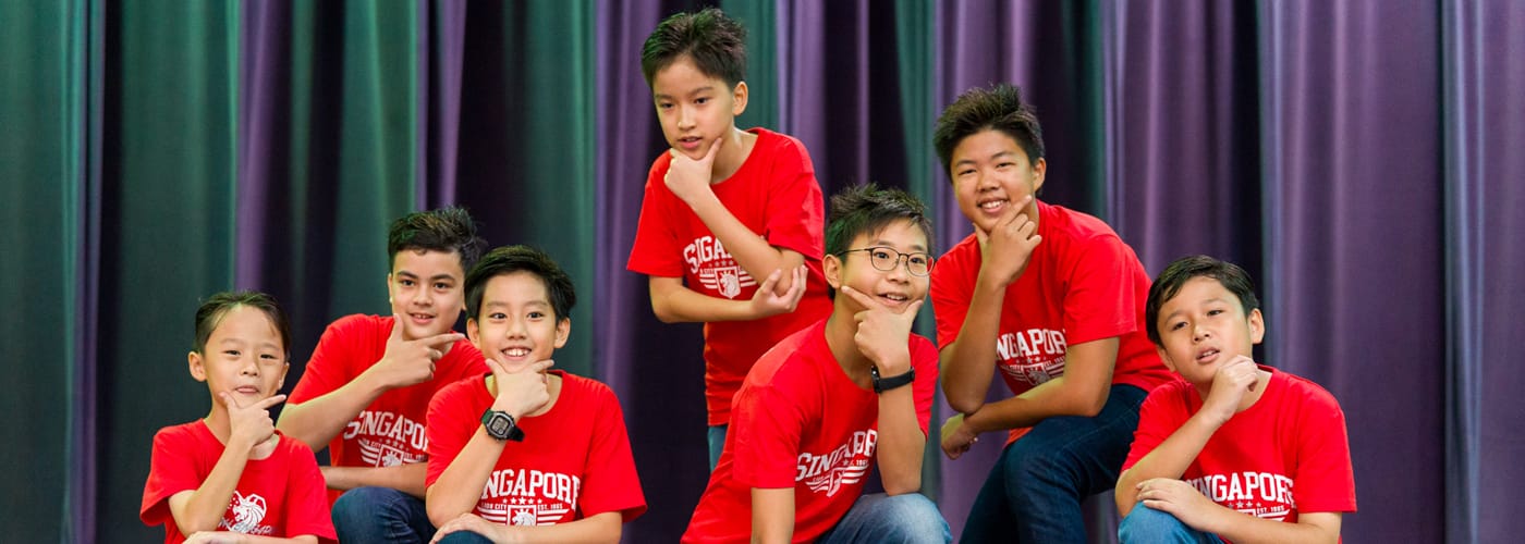 Seven boys in red Singapore shirts pose together with chin-resting hand gestures in front of colorful curtains.