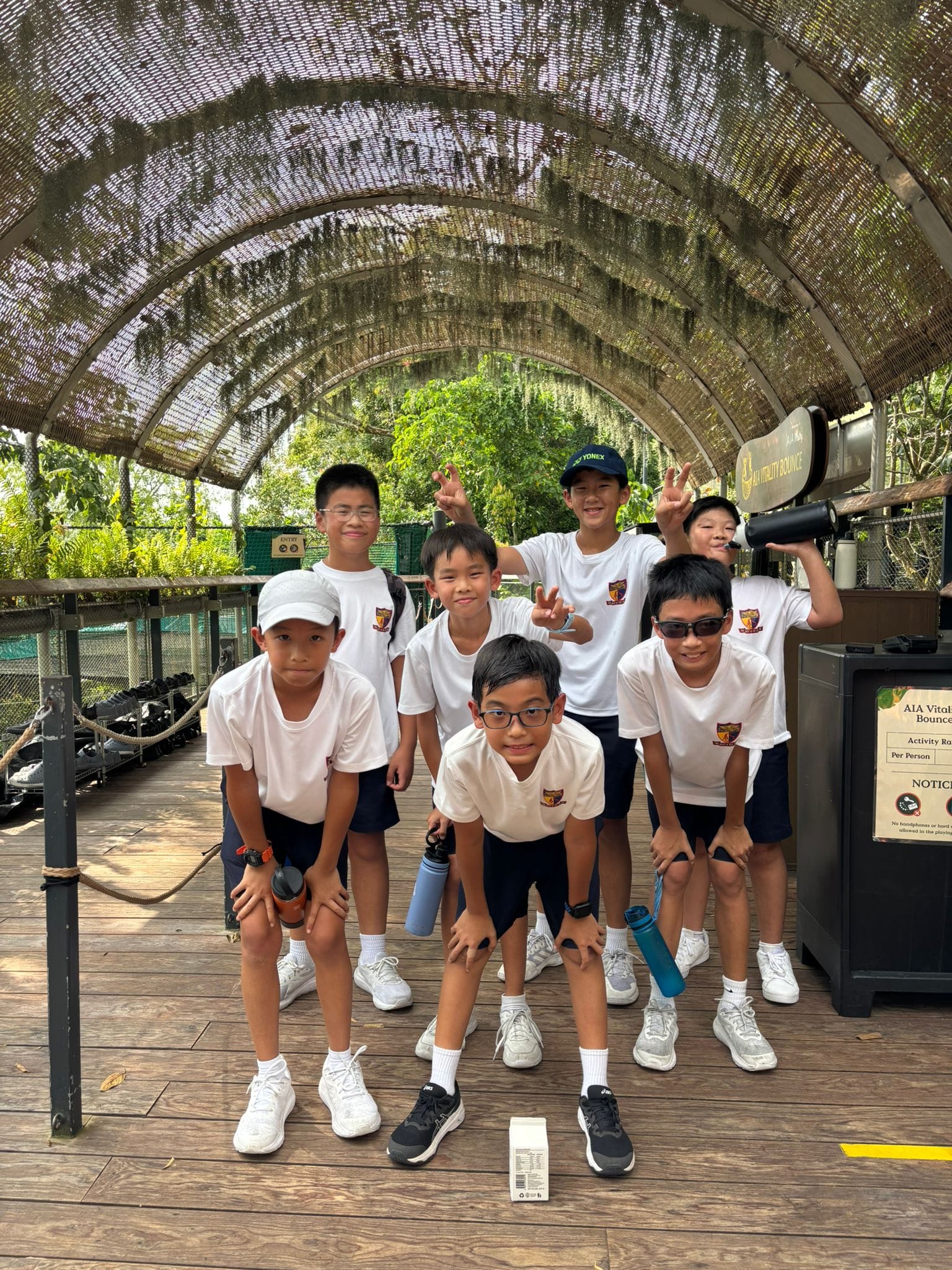 Seven boys in white uniforms pose under an arched tunnel walkway surrounded by trees, some making peace signs.
