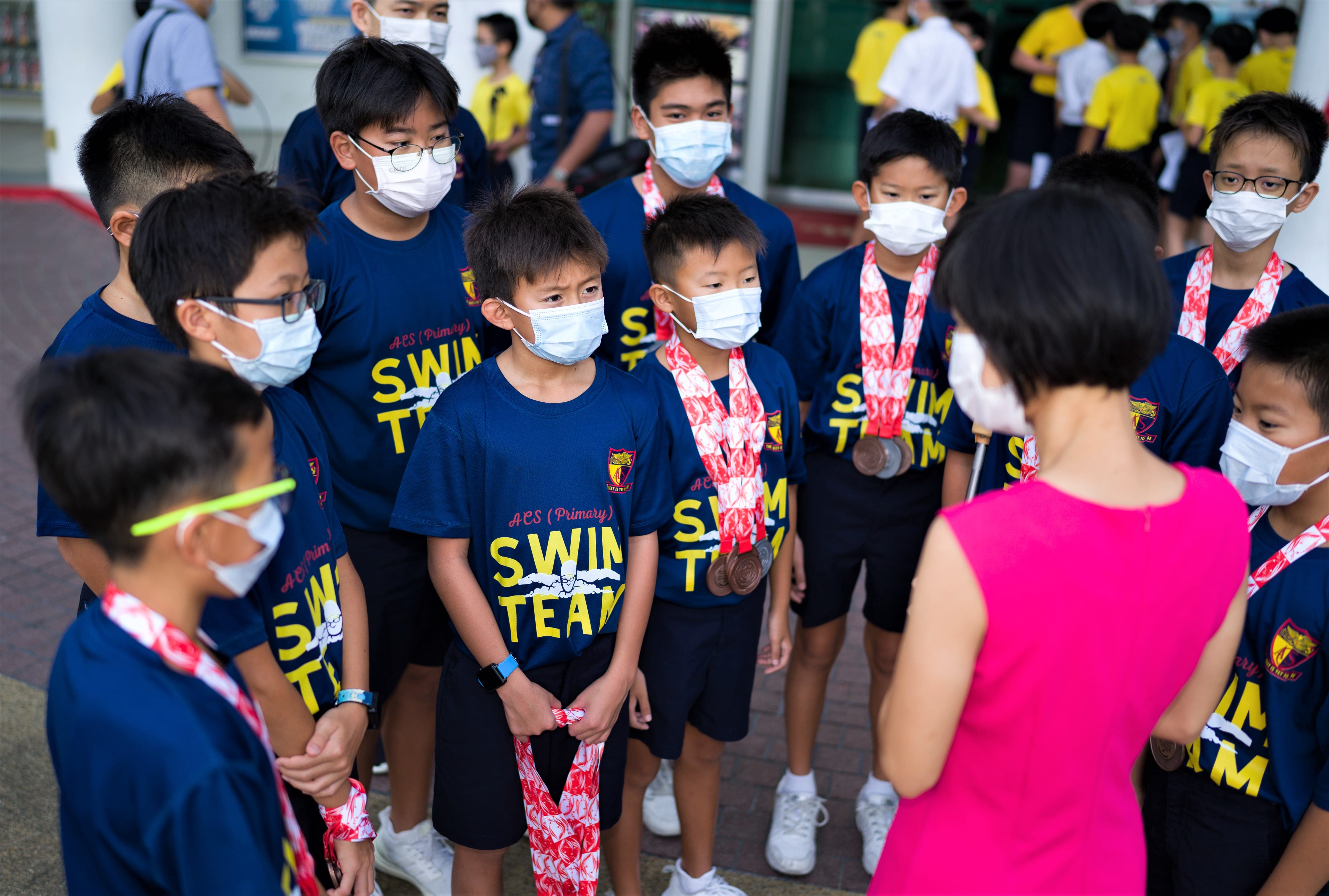 A group of masked boys in ACS Primary Swim Team shirts, some wearing medals, stand before a woman in pink.