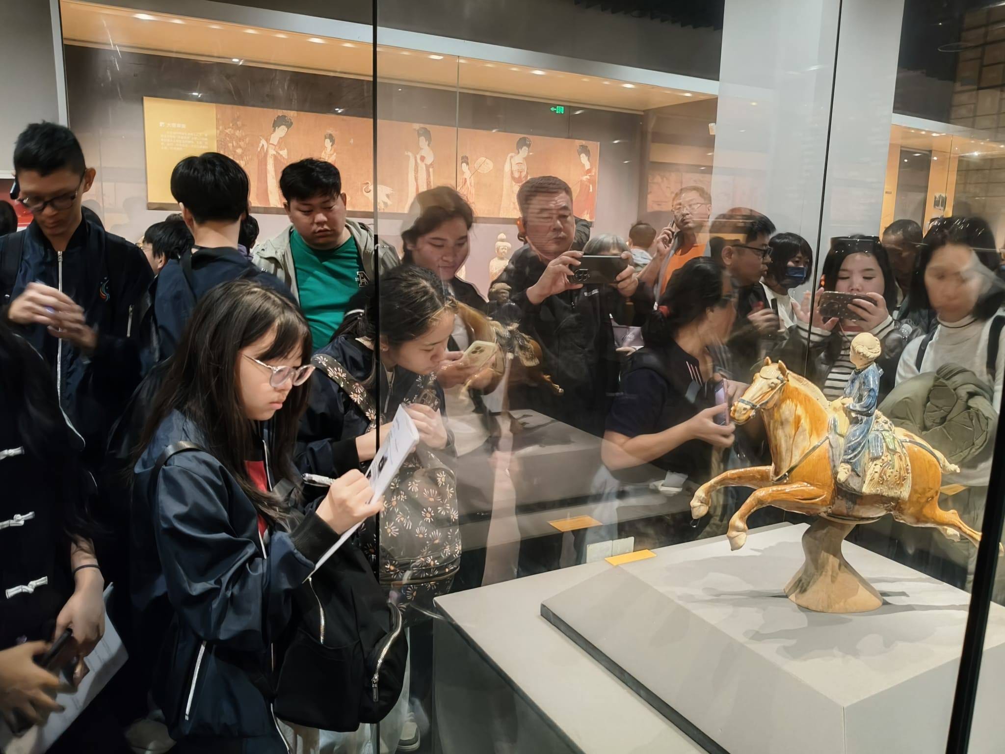 Crowd of museum visitors photographing and sketching a glazed ceramic horse-and-rider figurine in a glass display case.