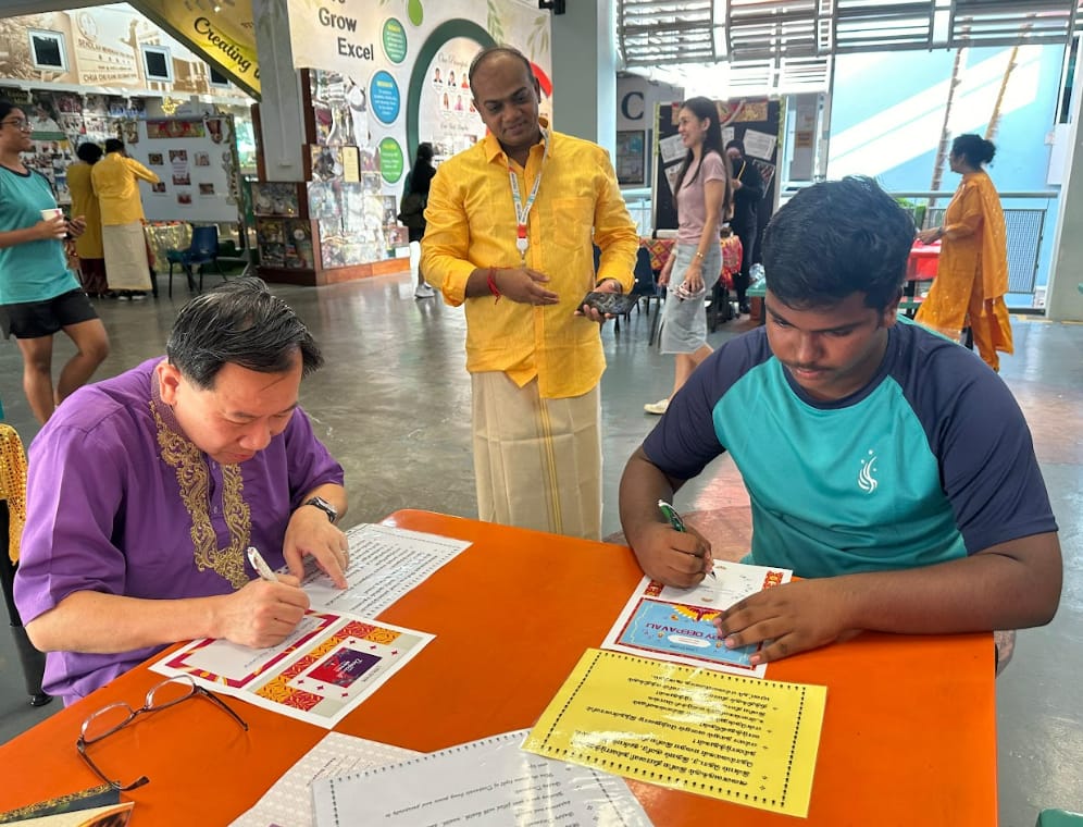 Two men writing on papers at an orange table during a cultural event, with others in traditional attire nearby.