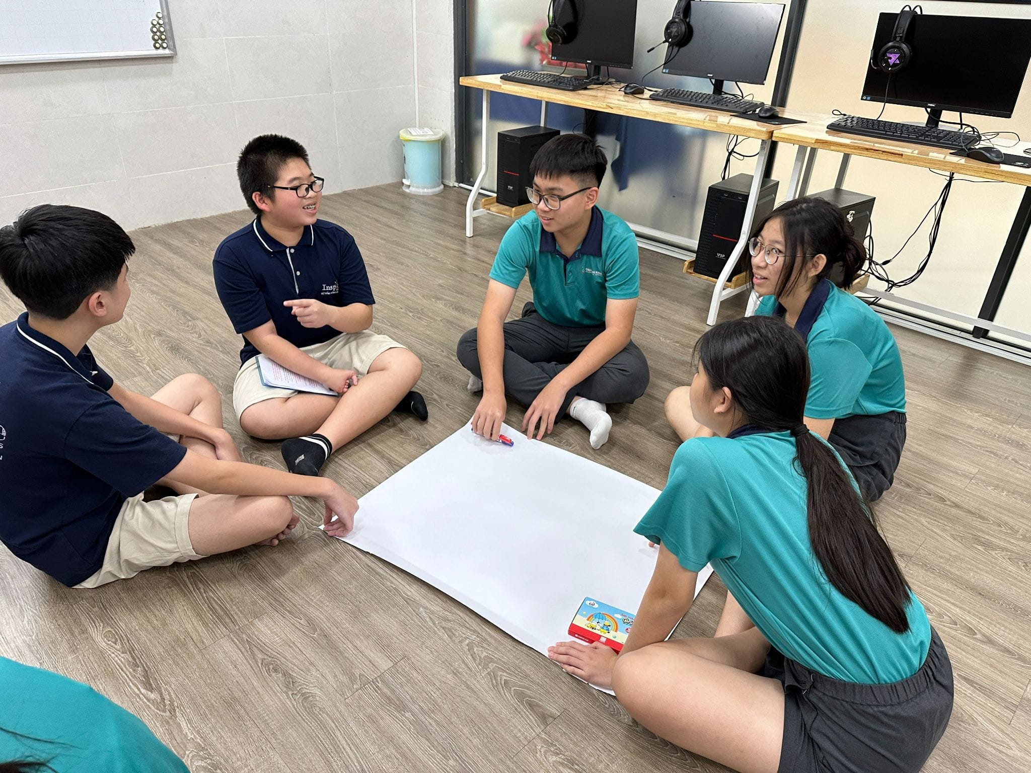 Five students in uniforms sit in a circle on a classroom floor, discussing over a large blank sheet of paper.
