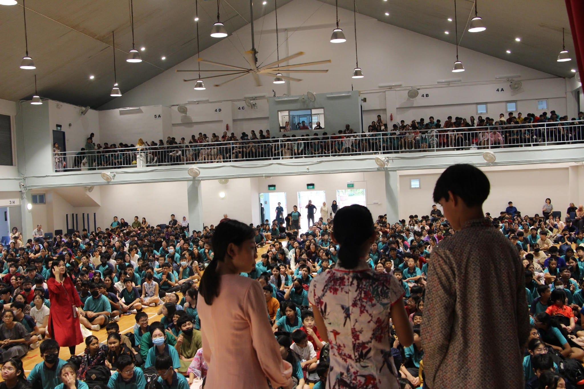 Three children stand on a stage facing a packed school hall filled with hundreds of students in teal uniforms.