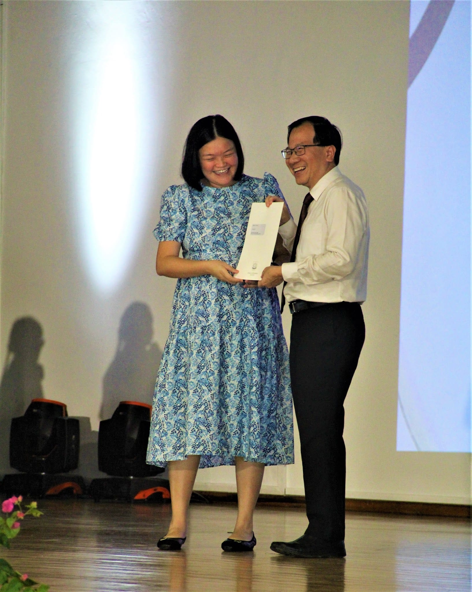 Two smiling people on a stage sharing a white envelope, woman in blue floral dress and man in white shirt and tie.