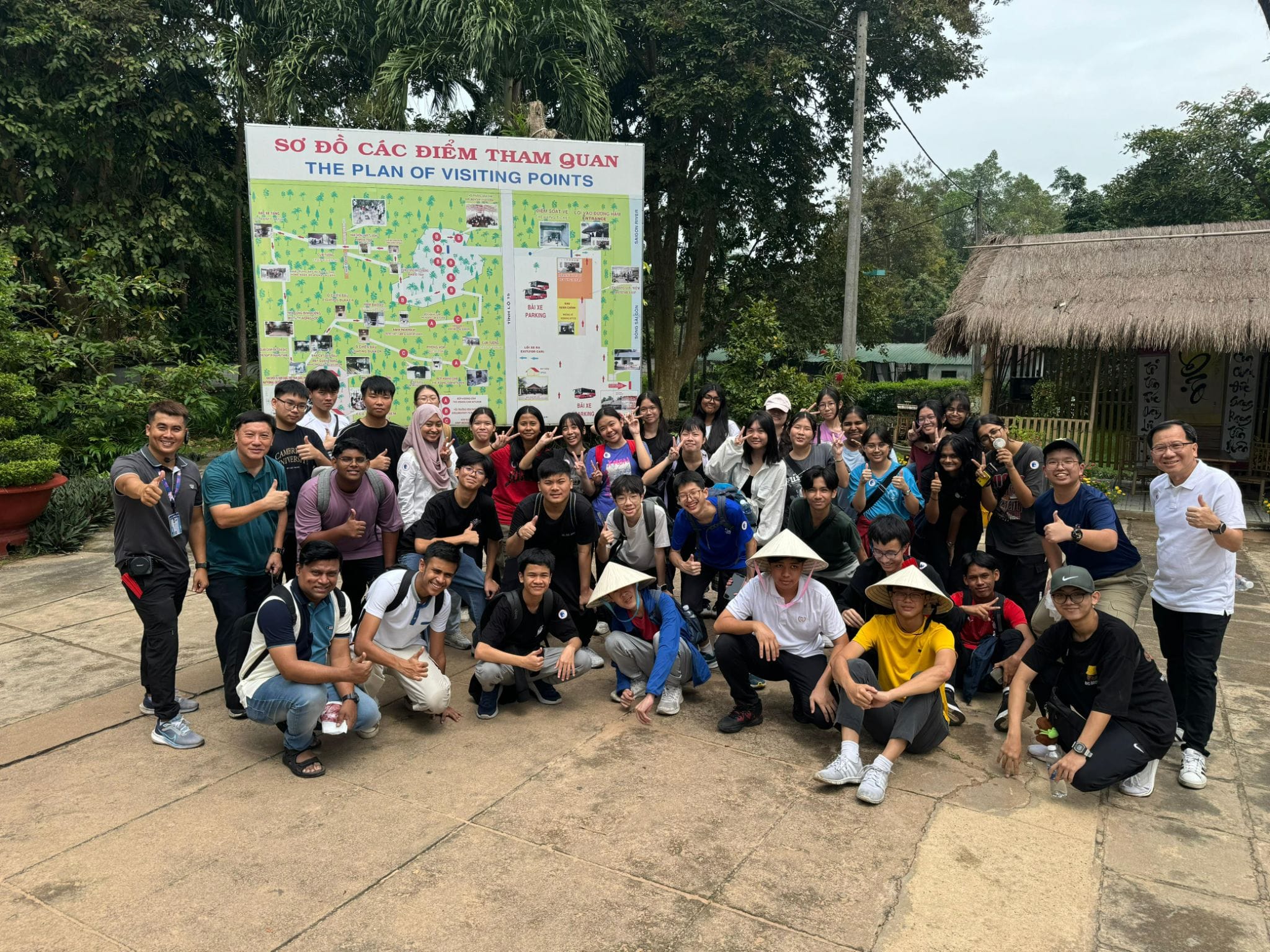 Large group of students posing in front of a Vietnamese visiting points map sign, some wearing traditional conical hats.