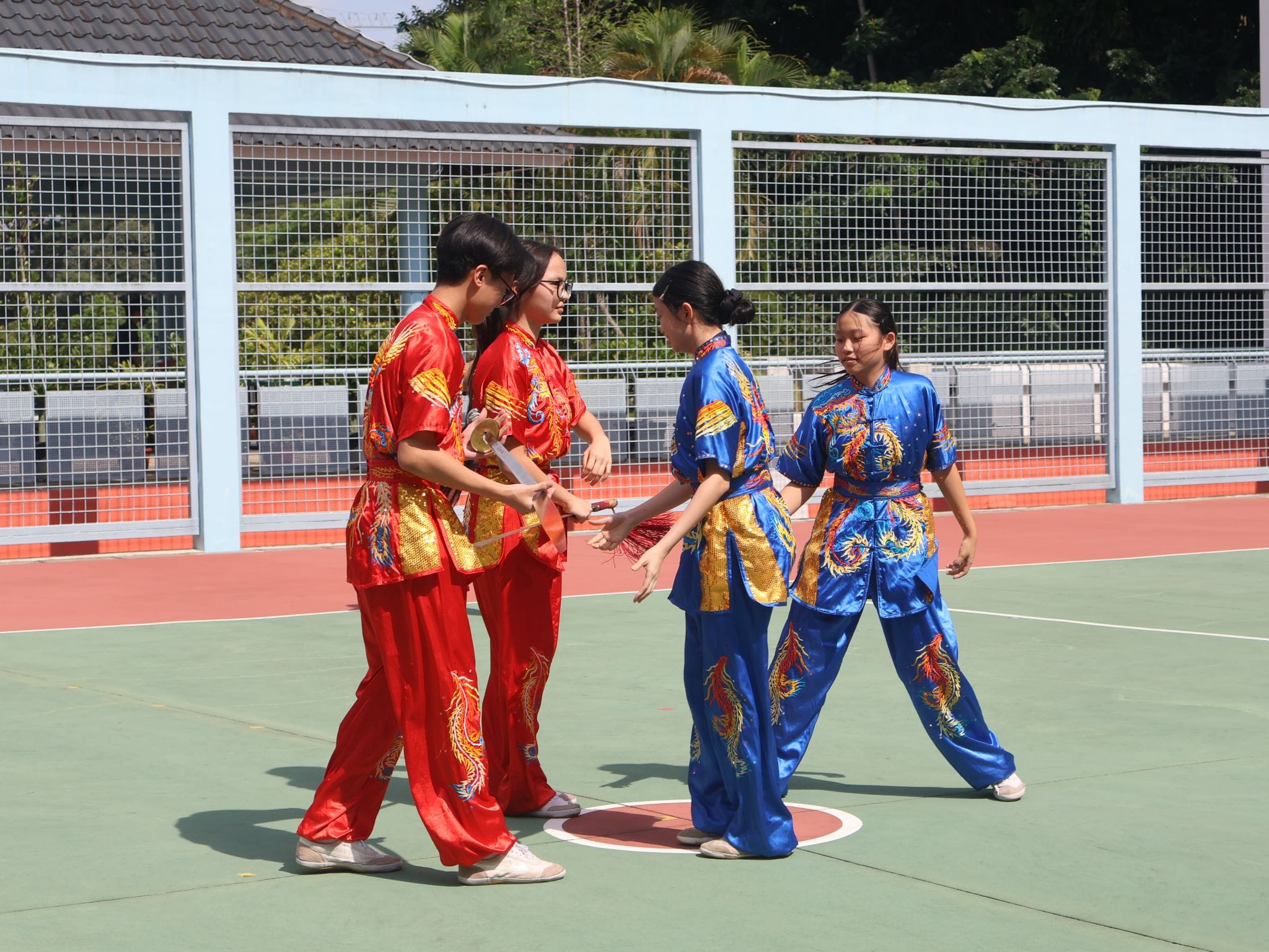 Four youth in red and blue dragon-embroidered wushu uniforms practicing sword moves on an outdoor court.