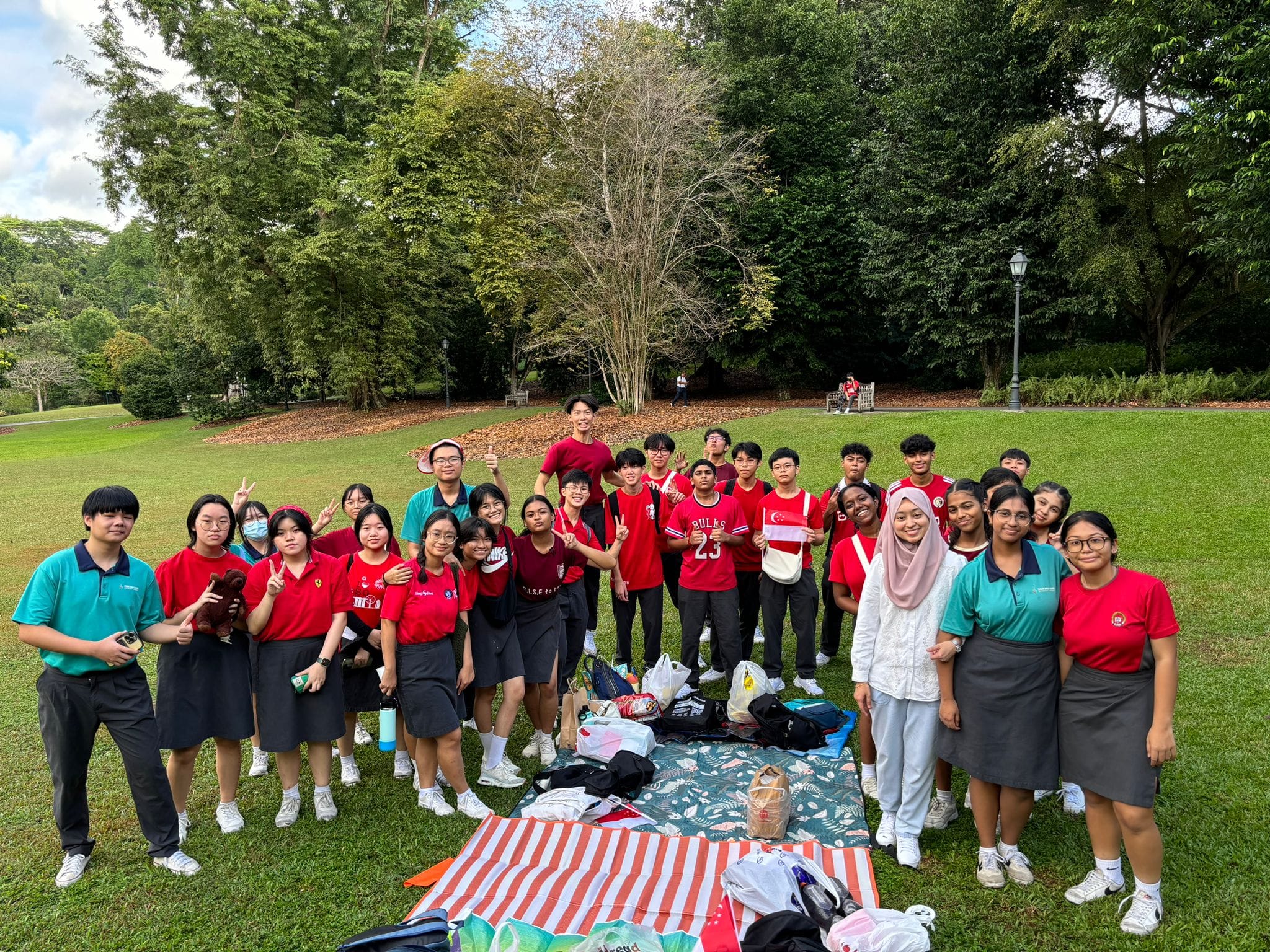 A large group of students in red and teal uniforms pose together on a grassy park lawn with picnic blankets nearby.