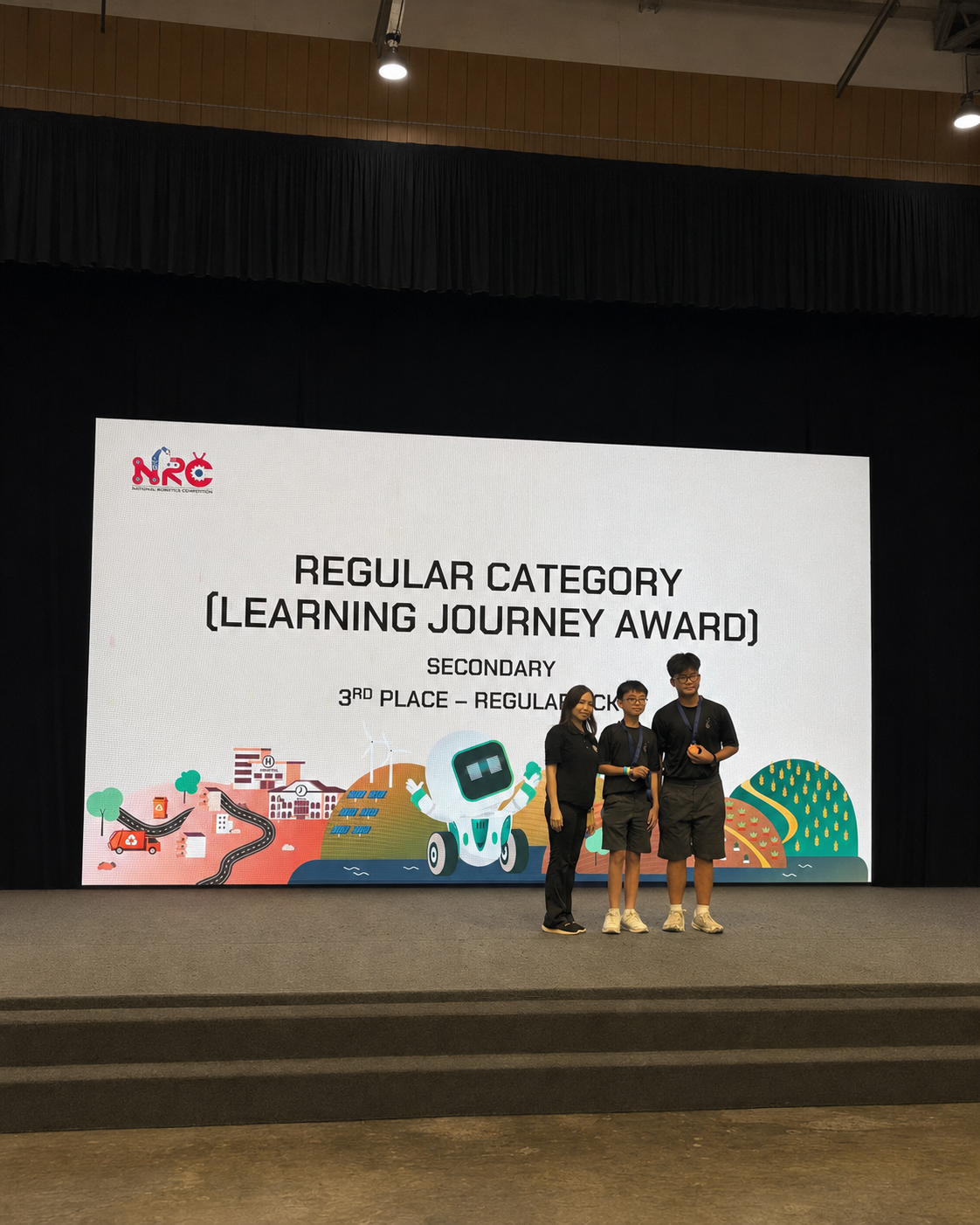 Three people stand on stage before an NRC screen announcing 3rd Place Regular Category Learning Journey Award.