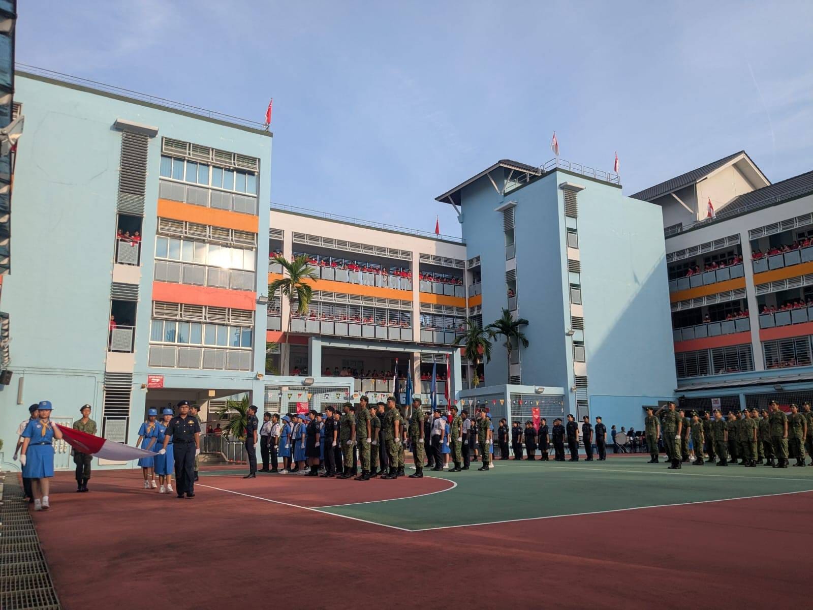 Students in uniform stand in formation during a flag ceremony at a school courtyard, with spectators watching from building corridors.