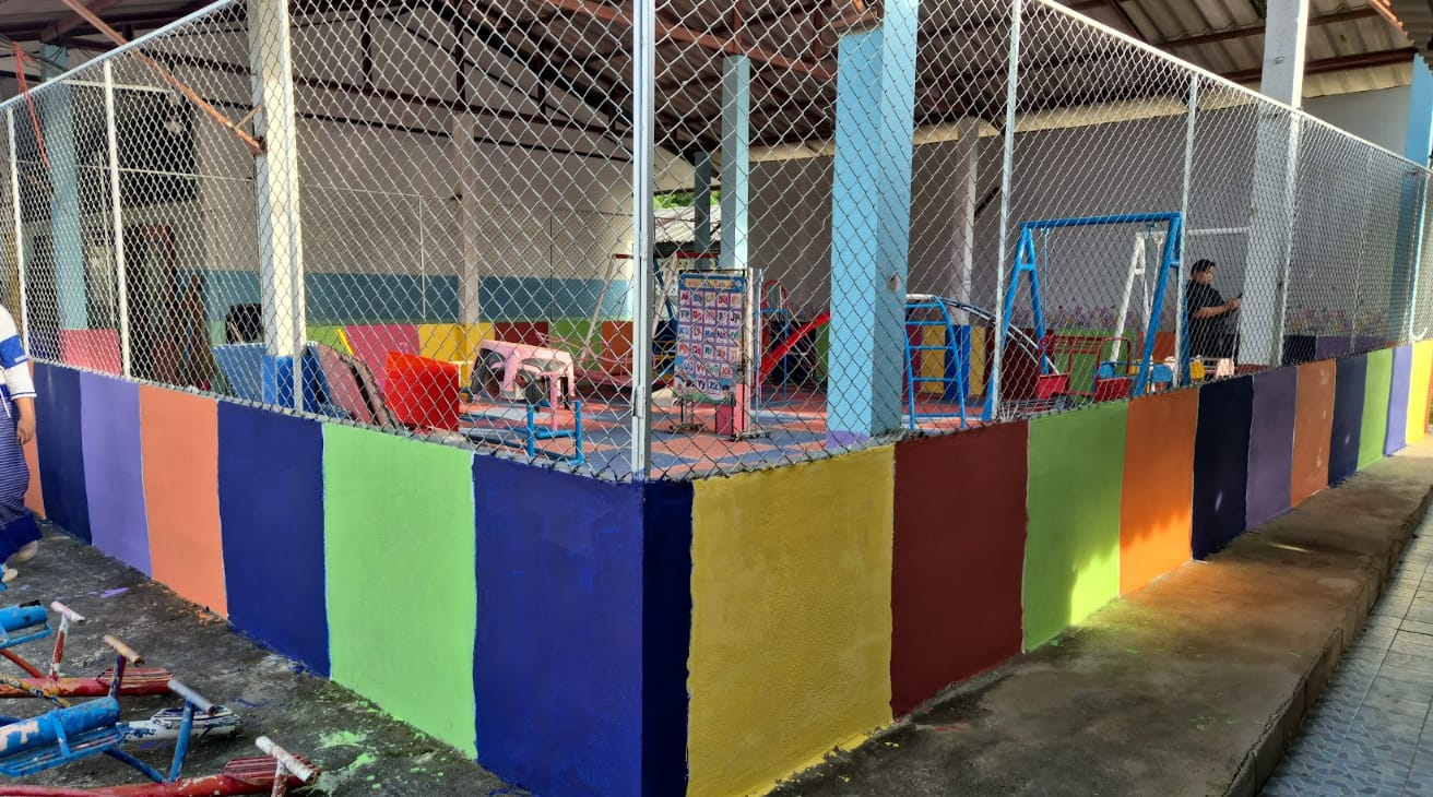 A fenced outdoor play area with a wall painted in multicolored vertical stripes and playground equipment inside.