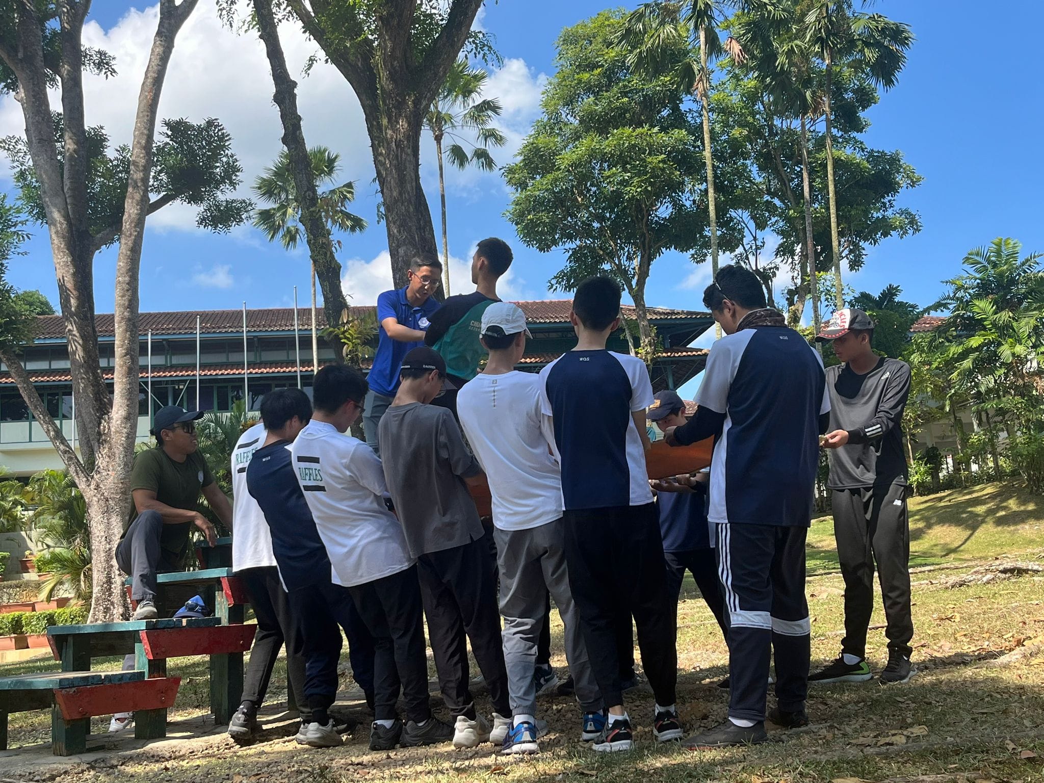 A group of young people gathered outdoors near trees and a building, huddled around an object on a sunny day.