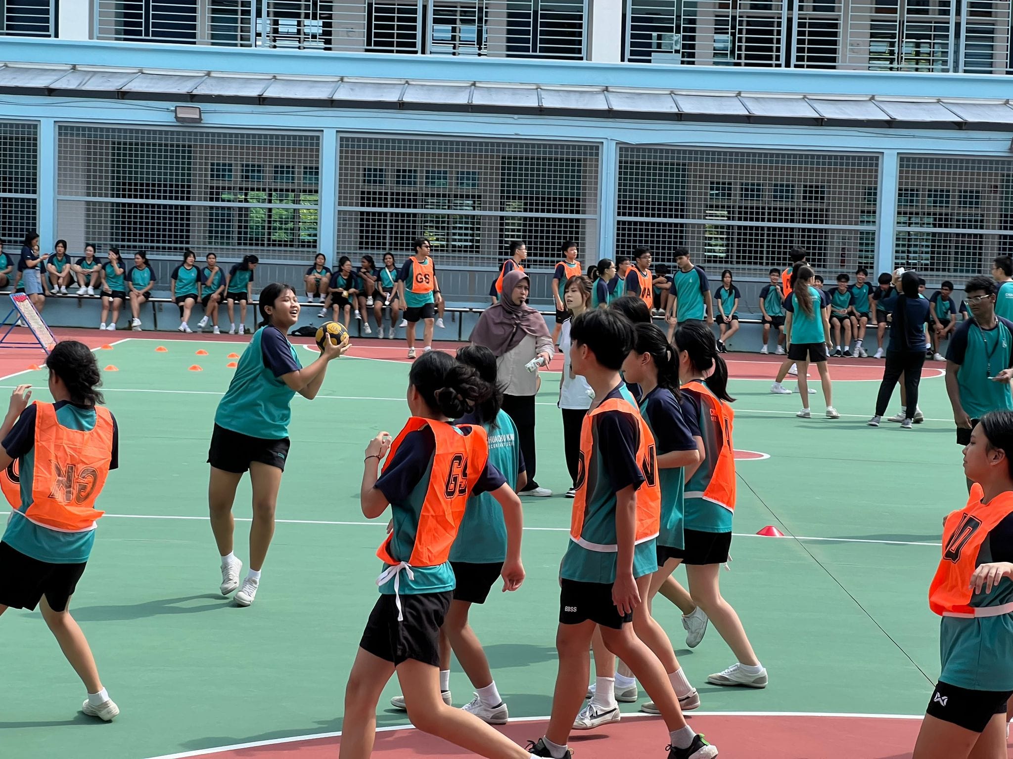 Students in teal shirts and orange vests play netball on an outdoor court while others watch from benches.