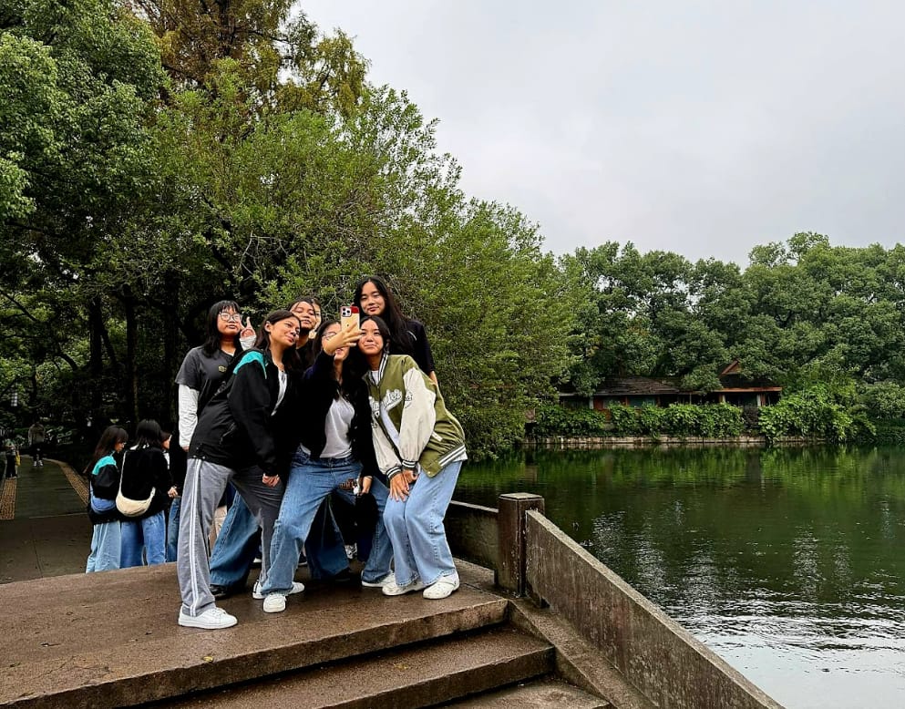 A group of young women take a selfie on stone steps beside a calm green pond surrounded by trees.