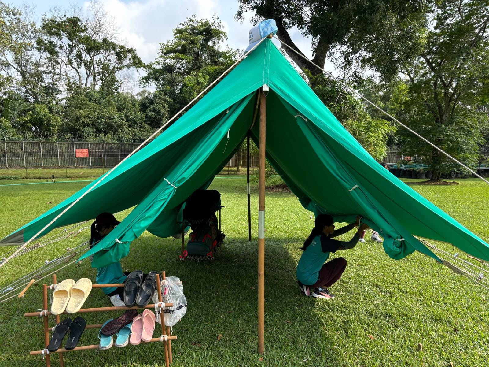 A green tarp tent on grass with two people inside, shoes on a bamboo rack in front, trees in background.