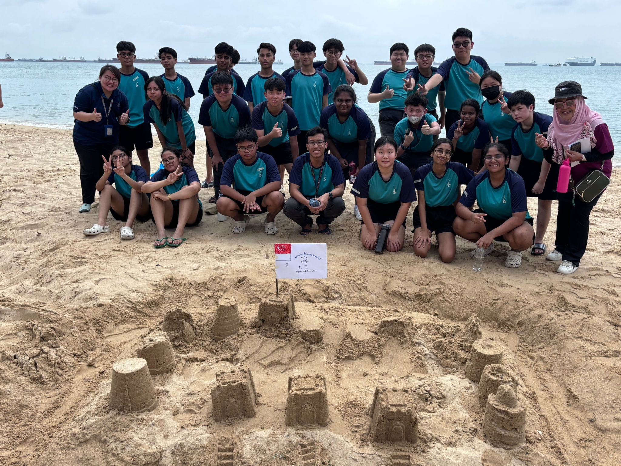 A large group of students in teal uniforms pose on a beach behind sand castles with a small Singapore flag.