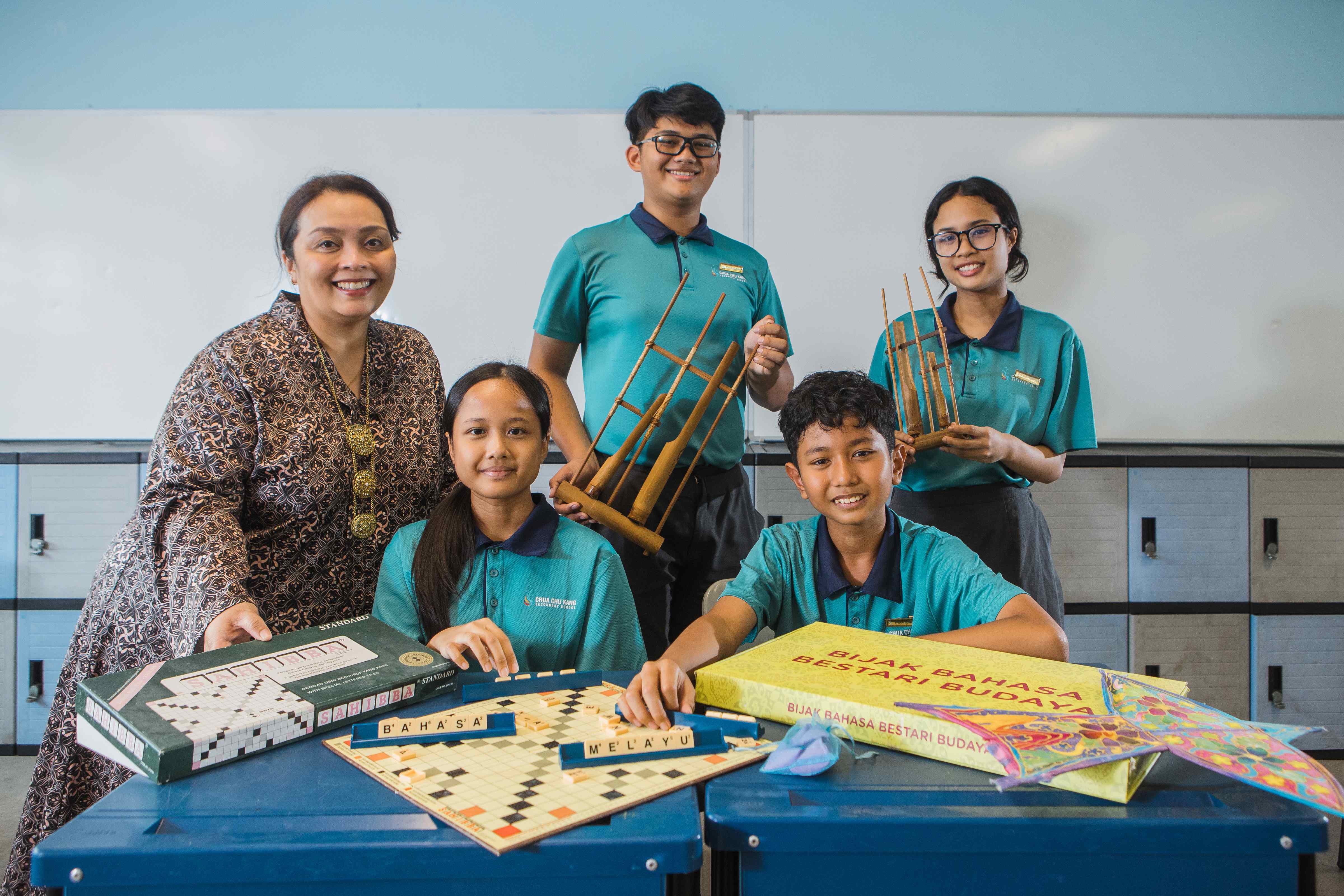 A teacher and four students in teal uniforms pose with a Scrabble board spelling Bahasa Melayu and traditional instruments.