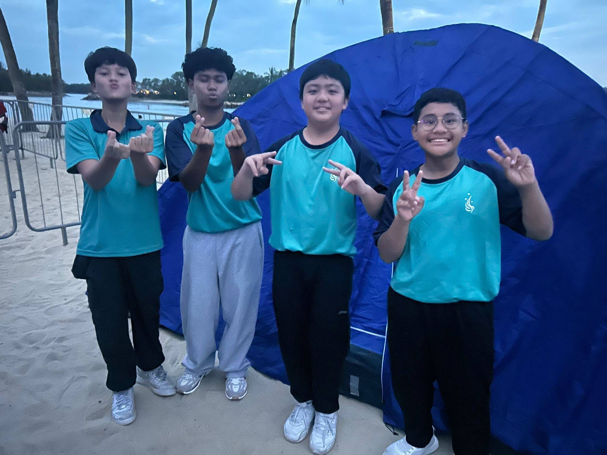 Four boys in matching teal uniforms pose together smiling near blue camping tents in an indoor venue.