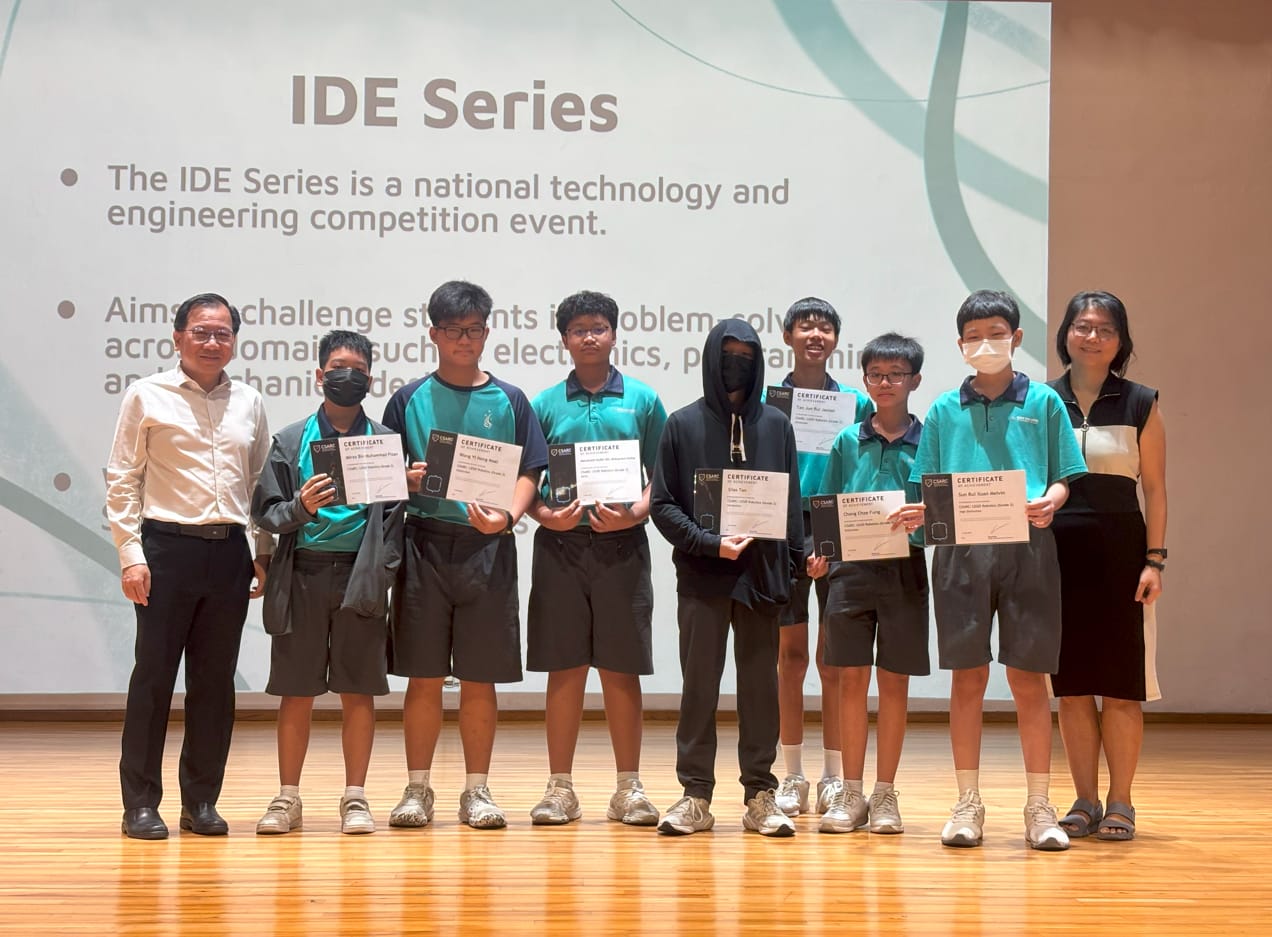 Students and two adults pose on a stage holding certificates in front of an IDE Series competition presentation slide.