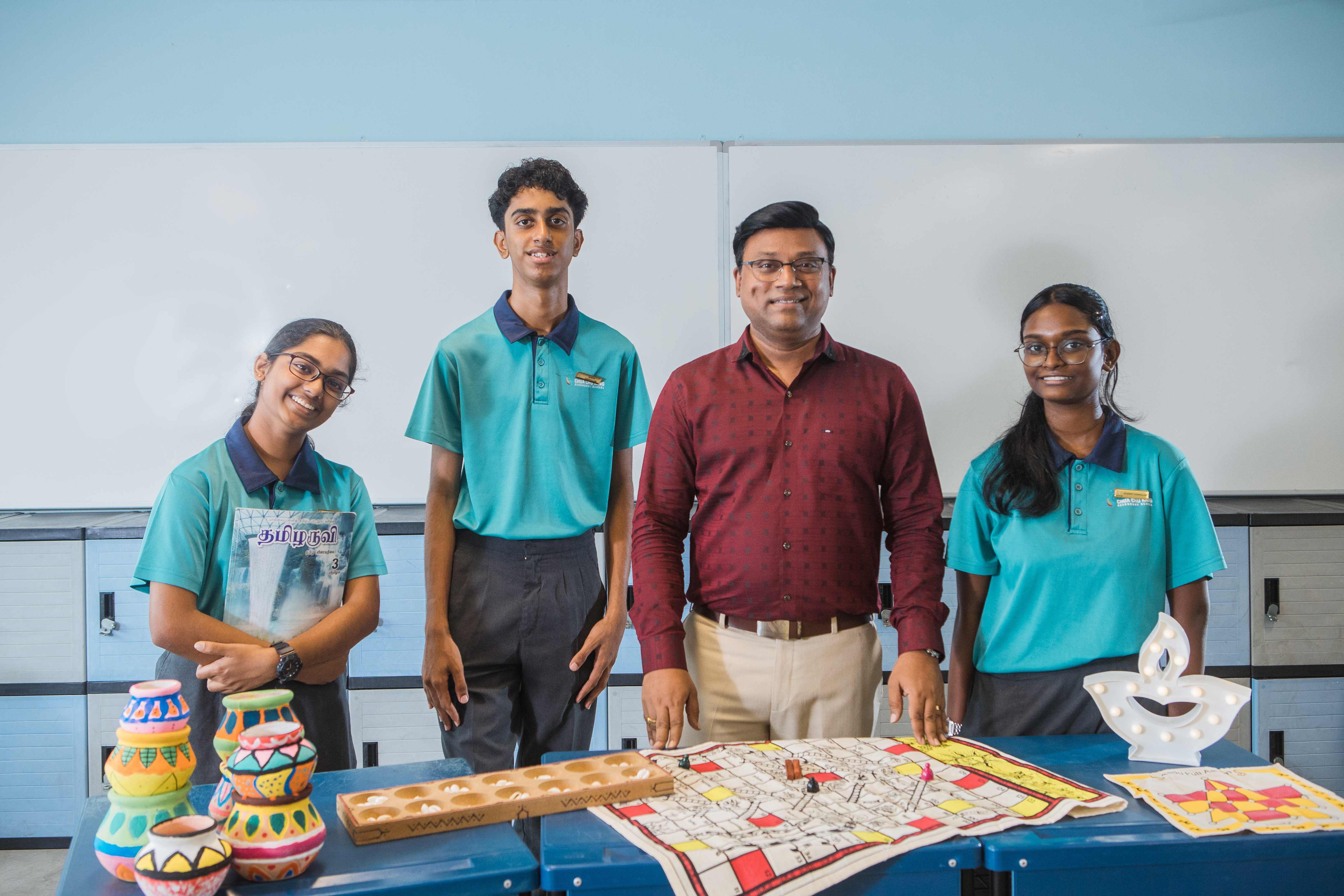Three students in teal uniforms and a teacher in a red shirt stand by a table with colorful pots and a board game.