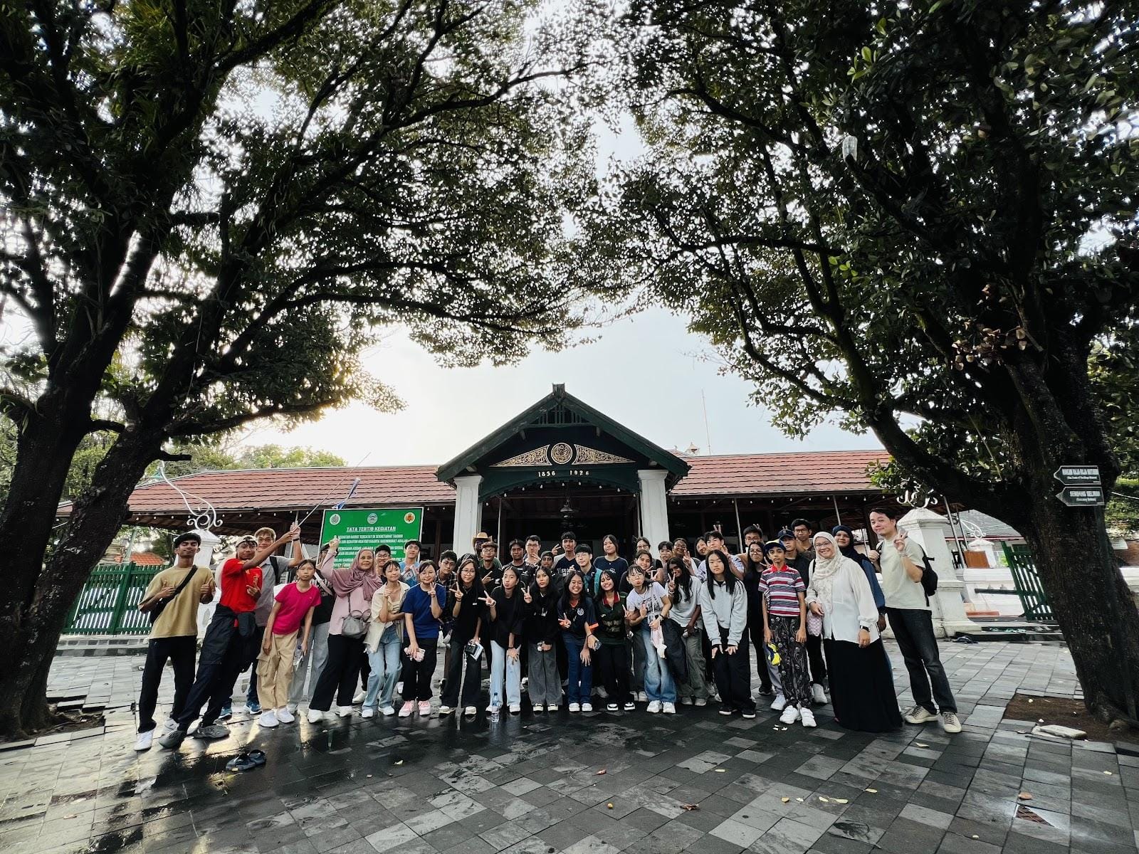 A large group of young people posing in front of a traditional Javanese pavilion surrounded by tall trees.