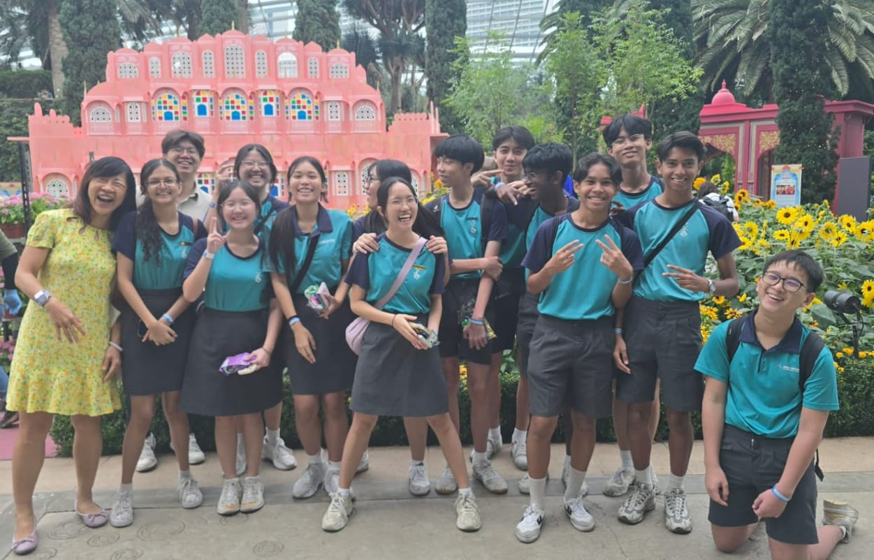 A group of smiling students in teal uniforms pose with teachers in front of a pink palace replica surrounded by sunflowers.