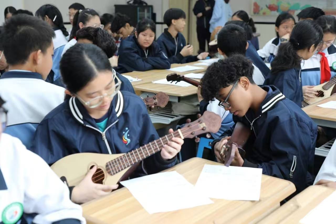 Students in navy uniforms sit at desks playing ukuleles in a classroom music lesson.