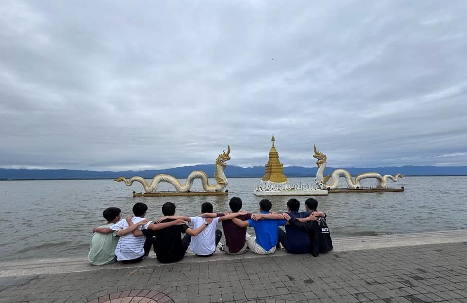 Seven people sit arm-in-arm by a lake facing two golden dragon statues flanking a small golden pagoda on water.