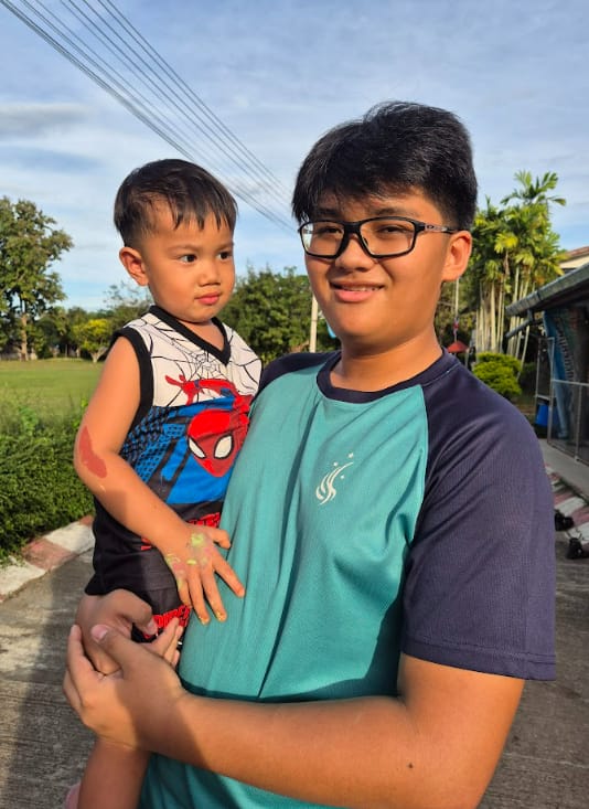 A teenage boy wearing glasses and a teal shirt holds a young toddler in a Spider-Man shirt outdoors.