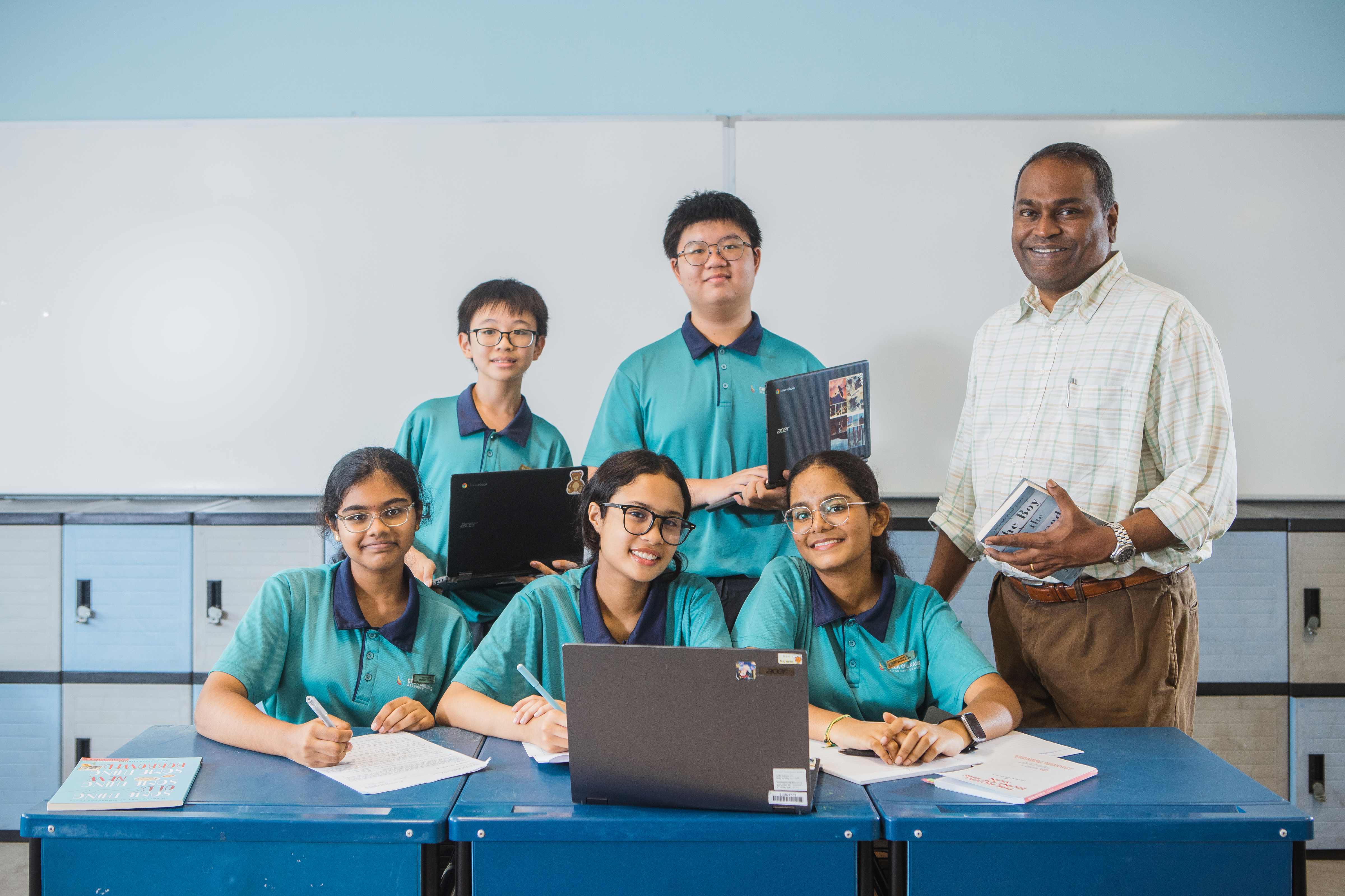 Five students in teal uniforms and a male teacher pose together in a classroom with laptops and books.