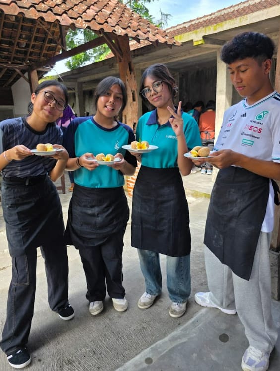 Four young people wearing black aprons hold white plates with small round food items outdoors near a building.