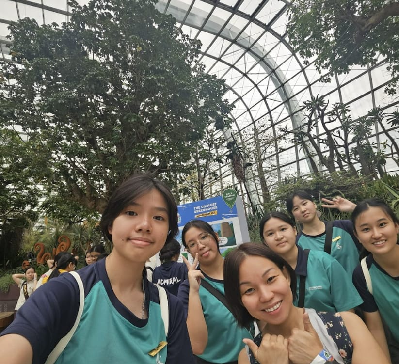 Six young women in teal uniforms take a selfie inside a large glass greenhouse with tall trees.