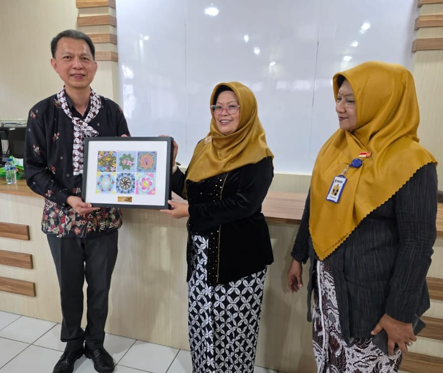 Three people stand together holding a framed collection of colorful mandala artworks indoors.