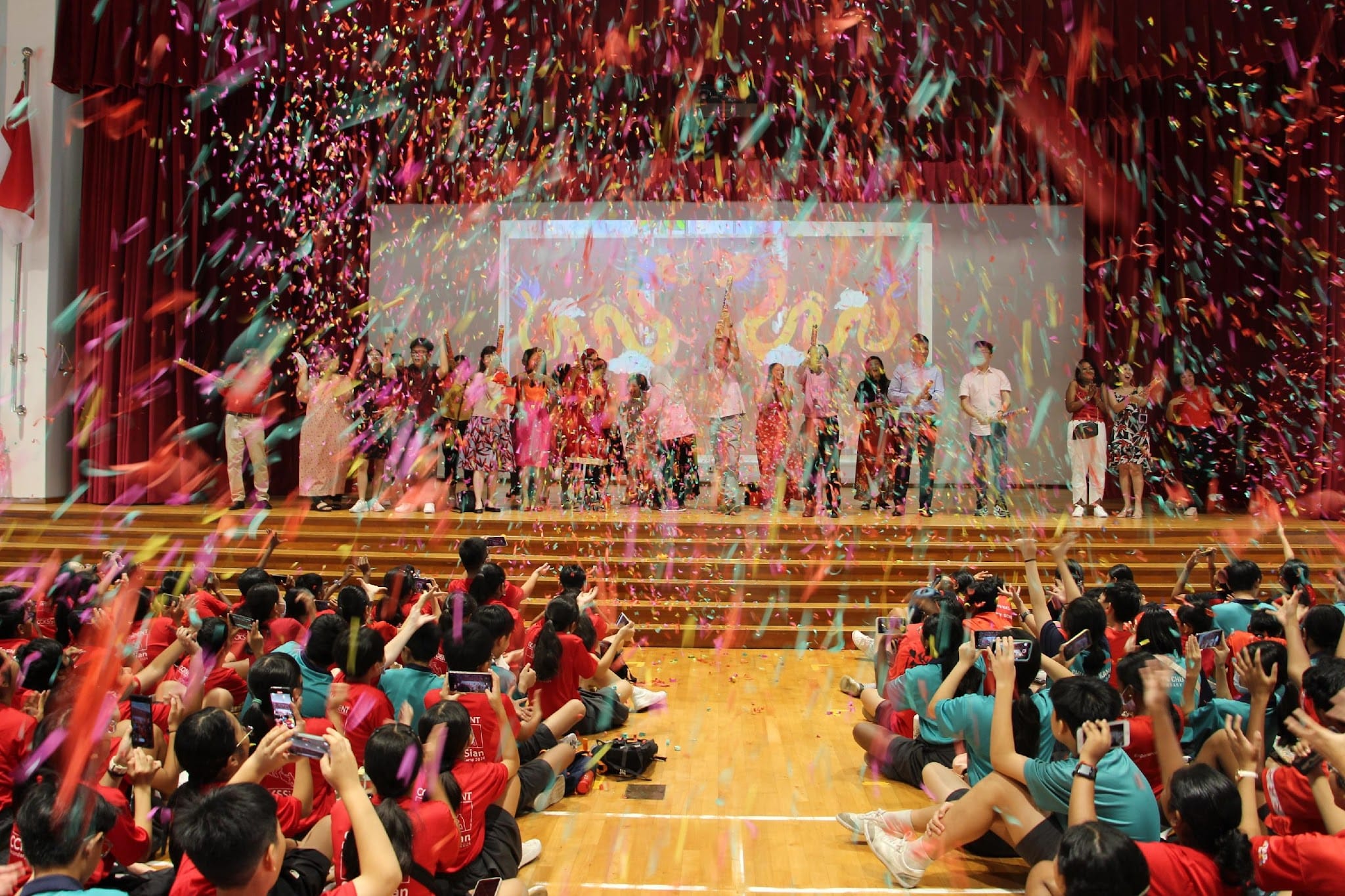 Students in red and teal shirts sit on a gym floor as colorful confetti fills the air during a stage celebration.