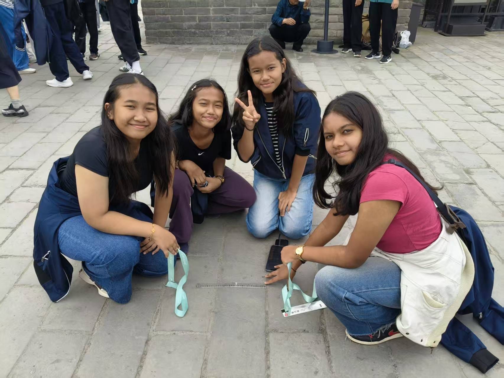 Four teenage girls crouch on a stone plaza, smiling at the camera, one making a peace sign.