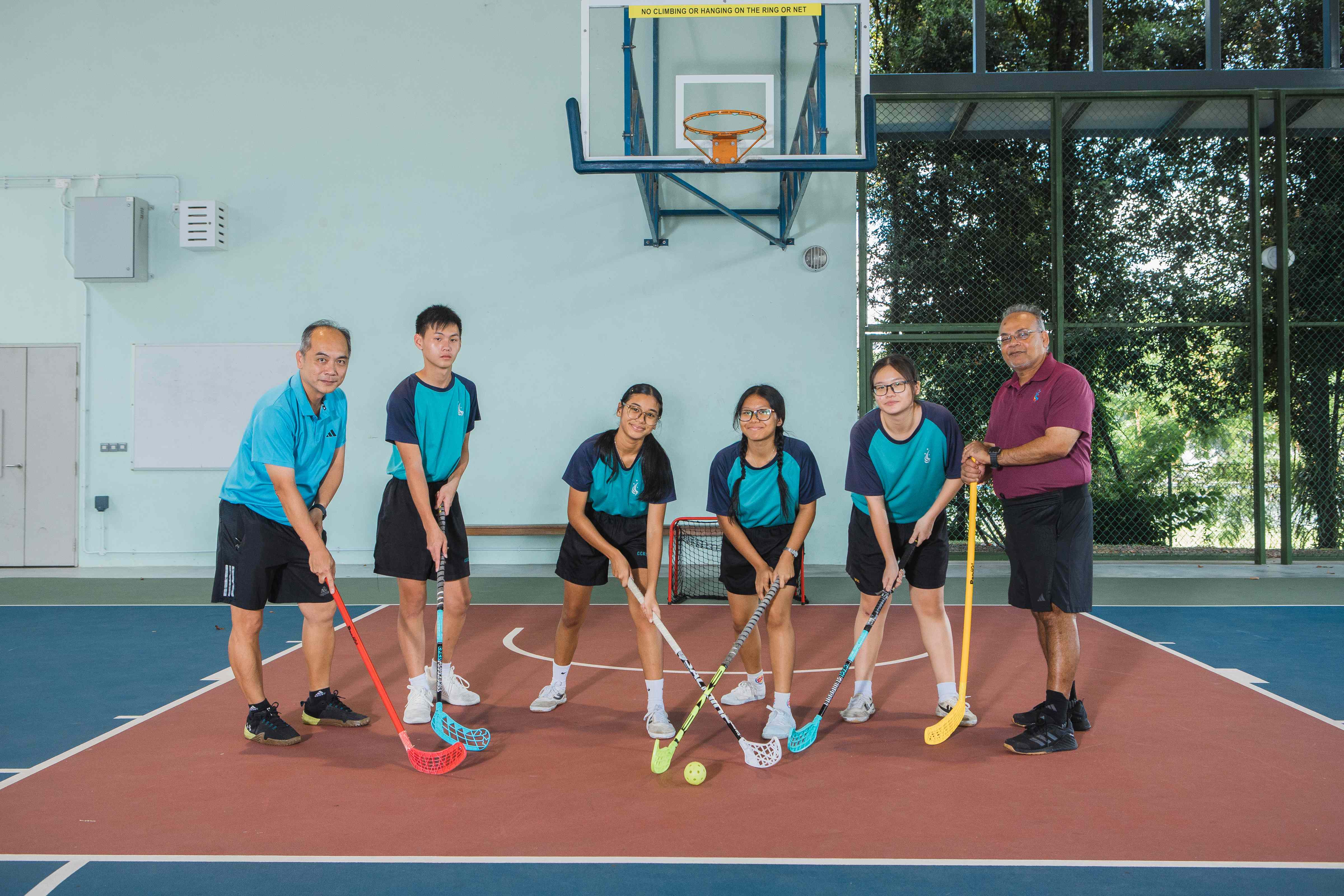 Six people holding floorball sticks pose on an indoor court with a basketball hoop on the wall behind them.