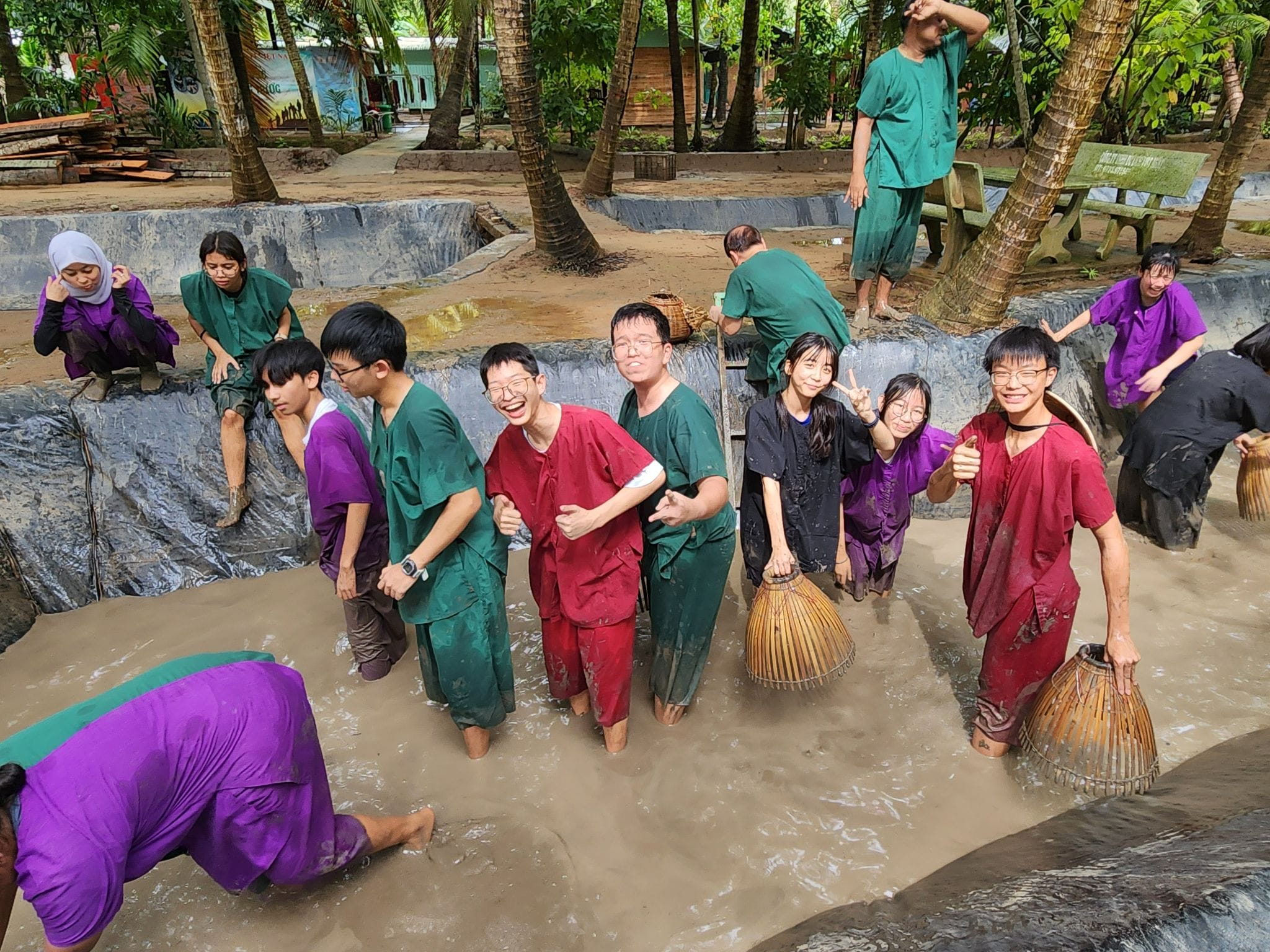 Group of people in colorful scrubs laughing and standing in a muddy outdoor pool surrounded by palm trees.