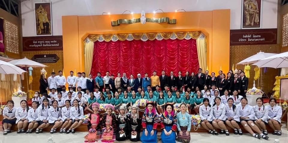 Large group of students and adults posing on a decorated stage, some wearing traditional Thai ethnic costumes.