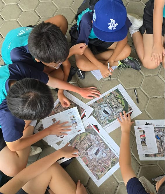 Several children sit on a paved ground, hudddling over printed maps and papers spread out between them.