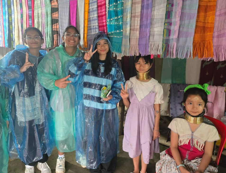 Five girls pose near colorful hanging scarves; three wear plastic rain ponchos, two wear traditional brass neck rings.