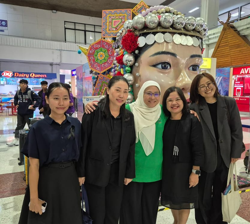 Five women pose in front of a large decorative face sculpture with an ornate headdress inside a shopping mall.
